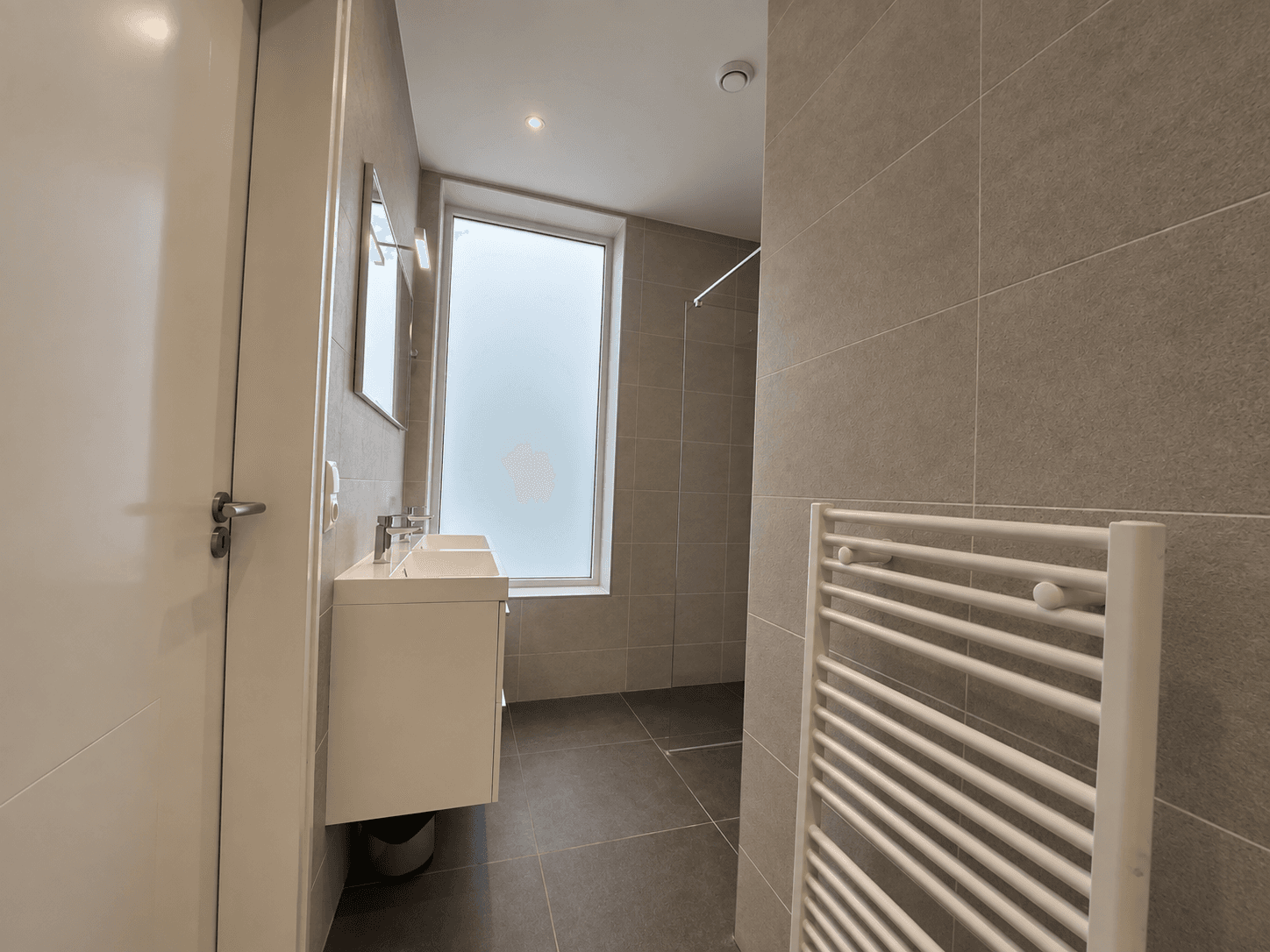Moderne badkamer met inloopdouche, wastafel en handdoekradiator, grote raampartij zorgt voor natuurlijk licht