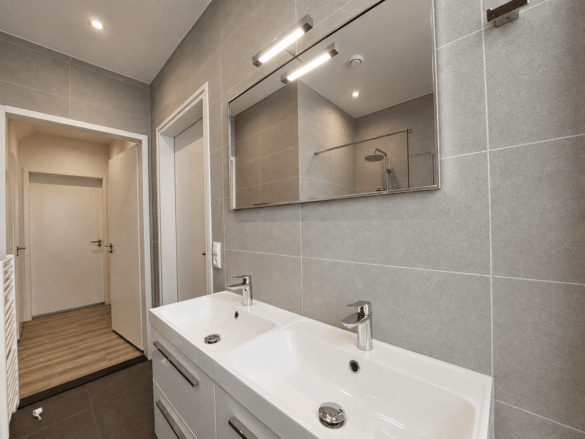 Moderne badkamer met dubbele wastafel, grote spiegel en douchecabine, grijze tegels en houten vloer in de gang.