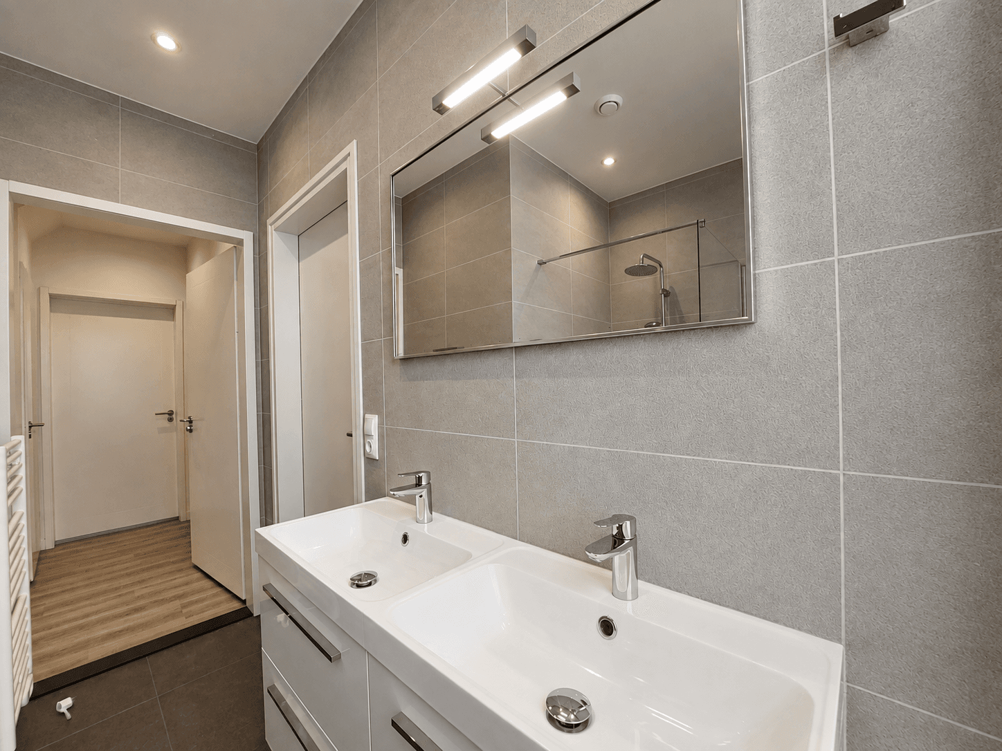 Moderne badkamer met dubbele wastafel, grote spiegel en douchecabine, grijze tegels en houten vloer in de gang.
