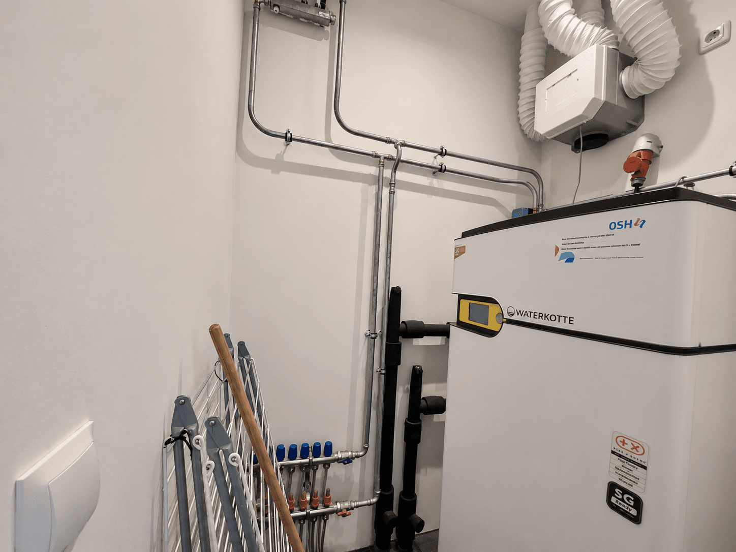 Technische ruimte met verwarmingsinstallatie en leidingen tegen de muur, inclusief een wasrek en ventilatiesysteem.