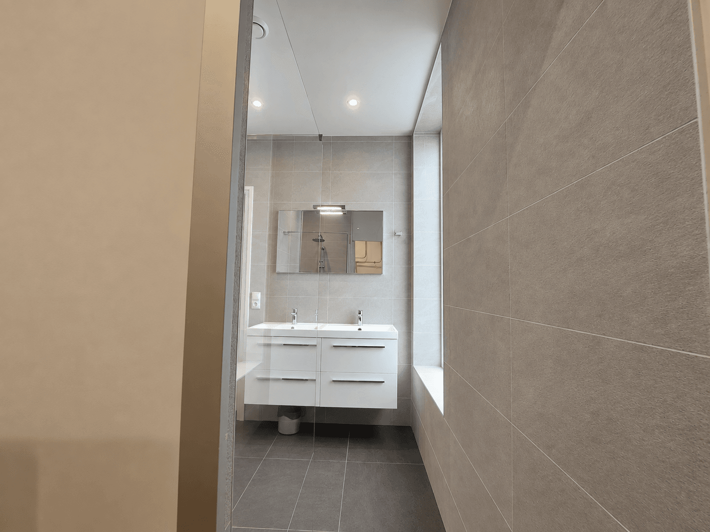 Moderne badkamer met grote wandtegels, dubbele wastafel en spiegel, goed verlicht en met zachte kleurtonen