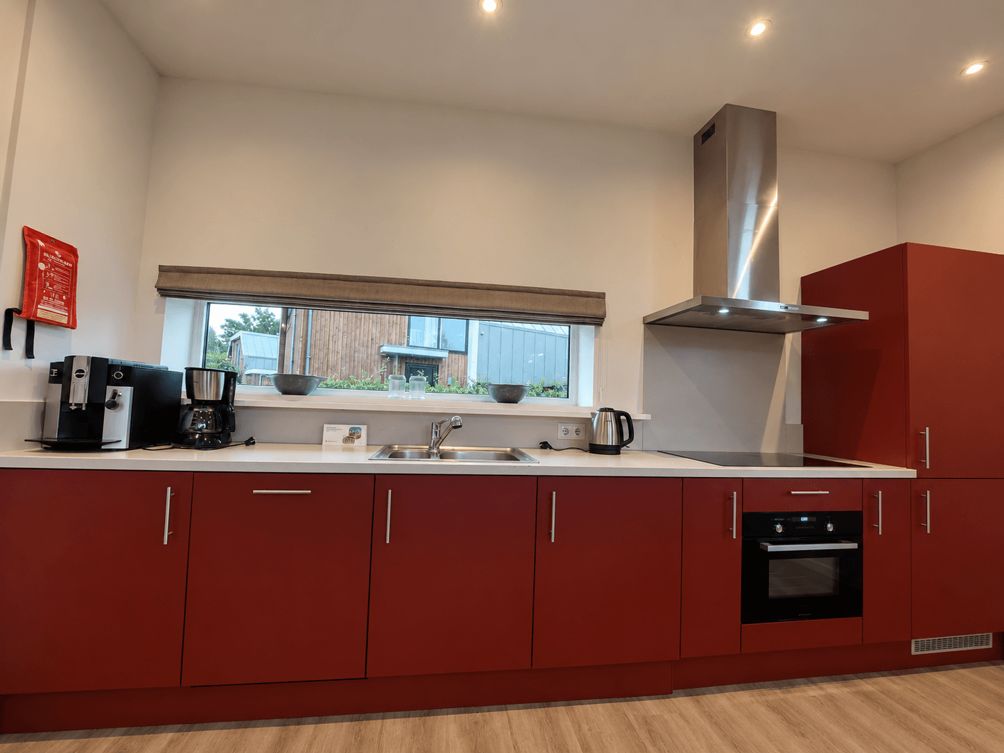 Moderne keuken met rode kastjes, kookplaat, afzuigkap en koffiezetapparaat aan het raam met uitzicht op buiten.