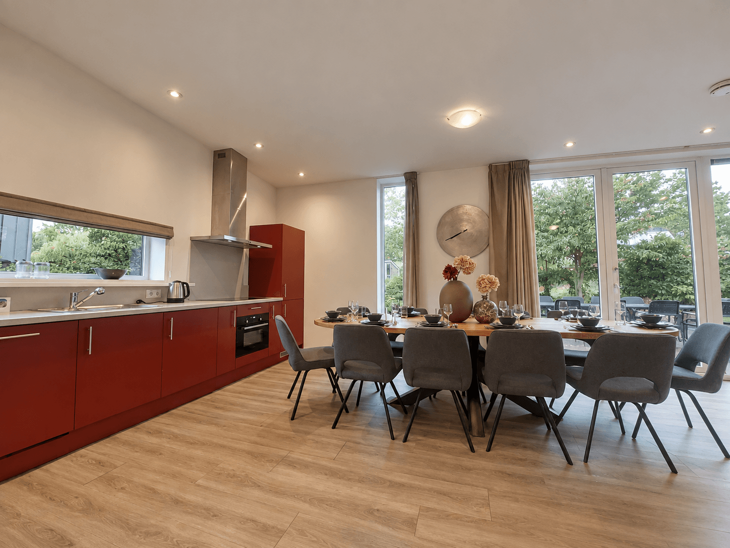 Moderne open keuken met rode kasten en een grote eettafel omringd door grijze stoelen, veel natuurlijk licht van grote ramen