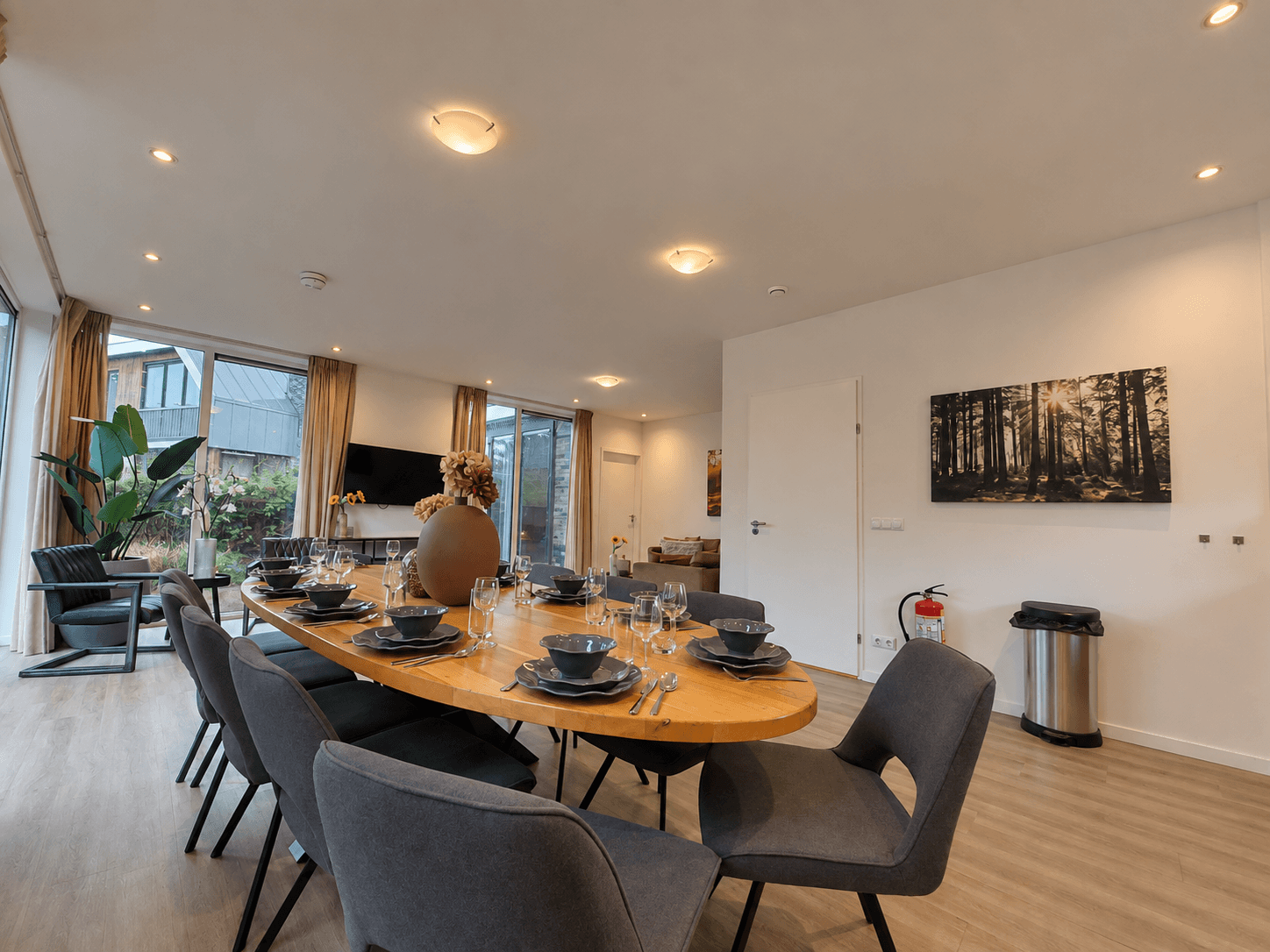 Eetkamer met grote tafel, zwarte stoelen, en ramen die uitzicht bieden op een groene buitenruimte.