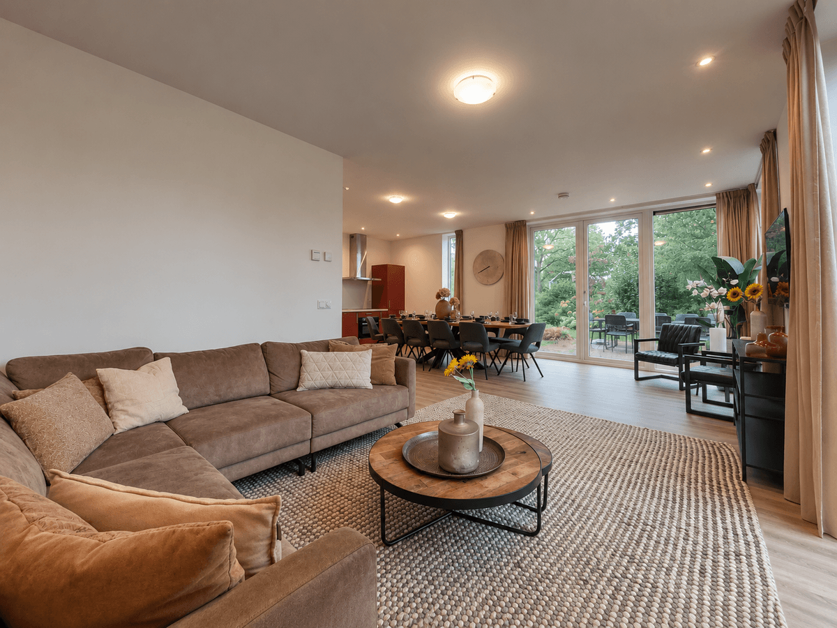 Woonkamer met een comfortabele loungebank, eettafel en grote ramen met uitzicht op de tuin.