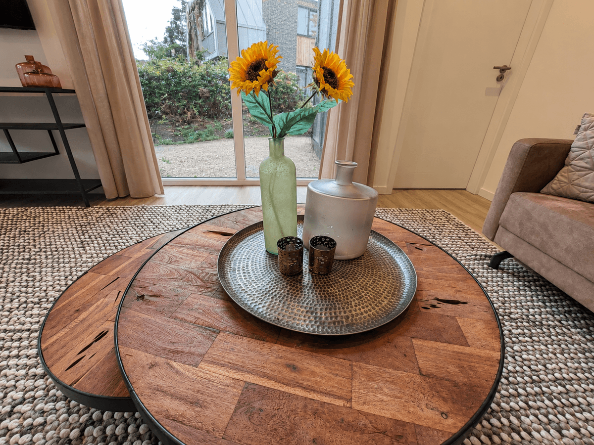 Koffietafel met een metalen schaal, bloemenvaas en decoratieve items voor een woonkamer met veel natuurlijk licht.