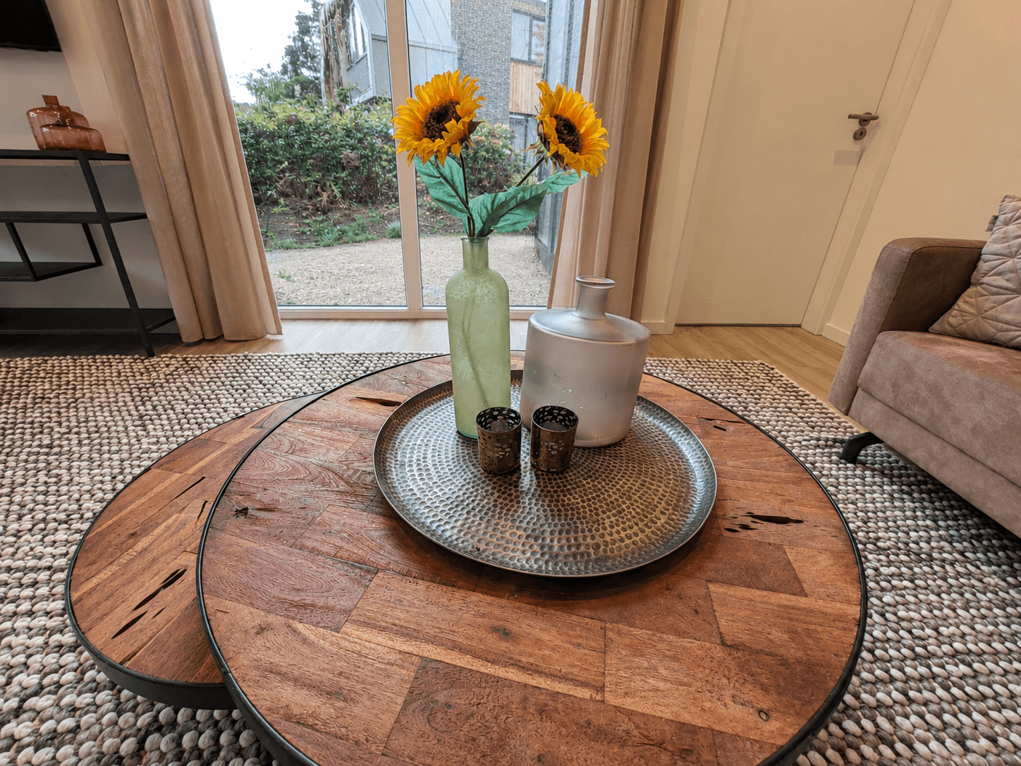 Koffietafel met een metalen schaal, bloemenvaas en decoratieve items voor een woonkamer met veel natuurlijk licht.