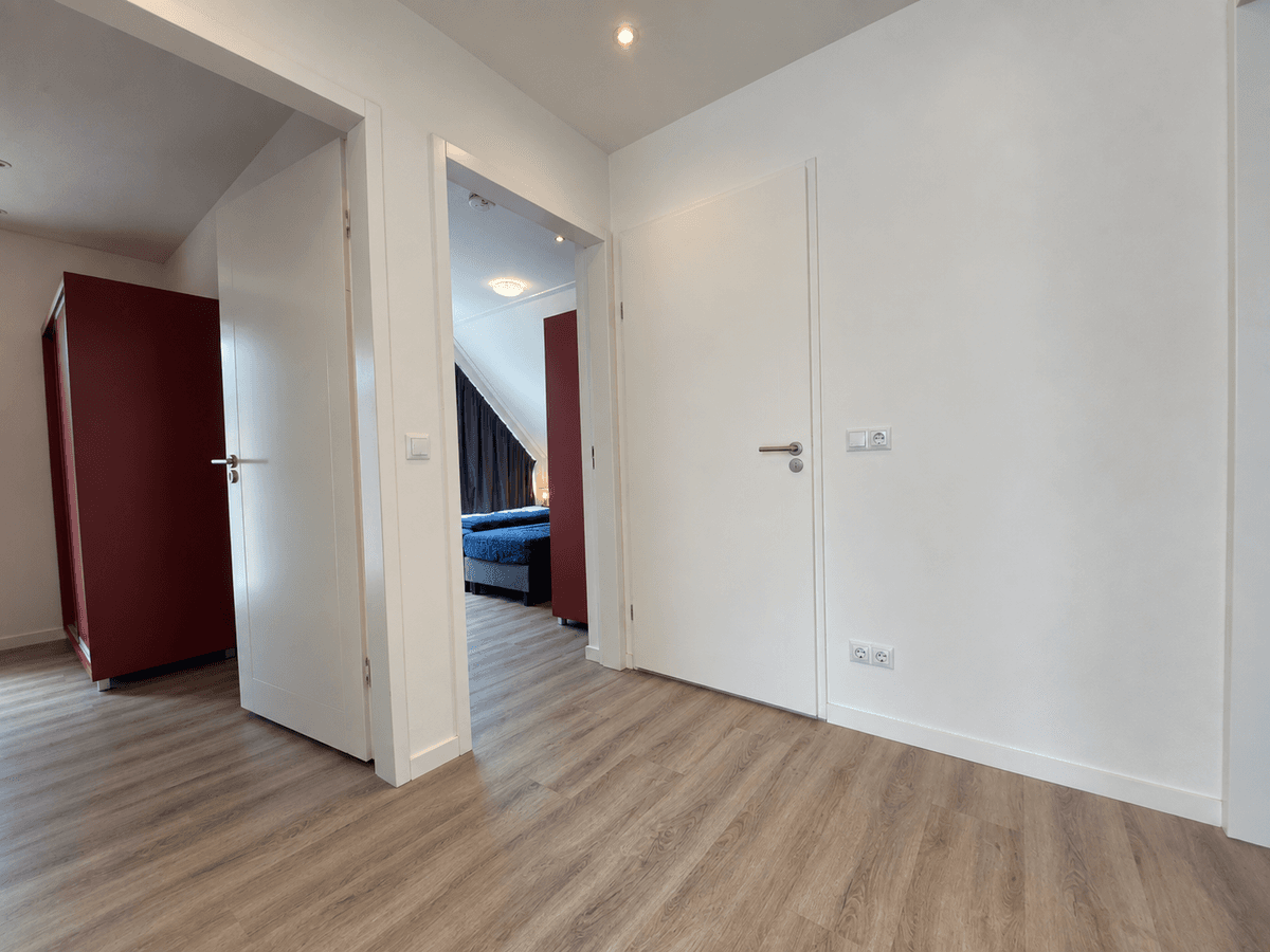 Gang met witte muren en licht houten vloeren, aan de rechterkant een slaapkamer met blauw bed en kastje, links een rode kast.