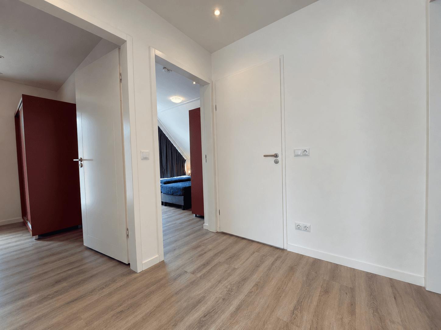Gang met witte muren en licht houten vloeren, aan de rechterkant een slaapkamer met blauw bed en kastje, links een rode kast.