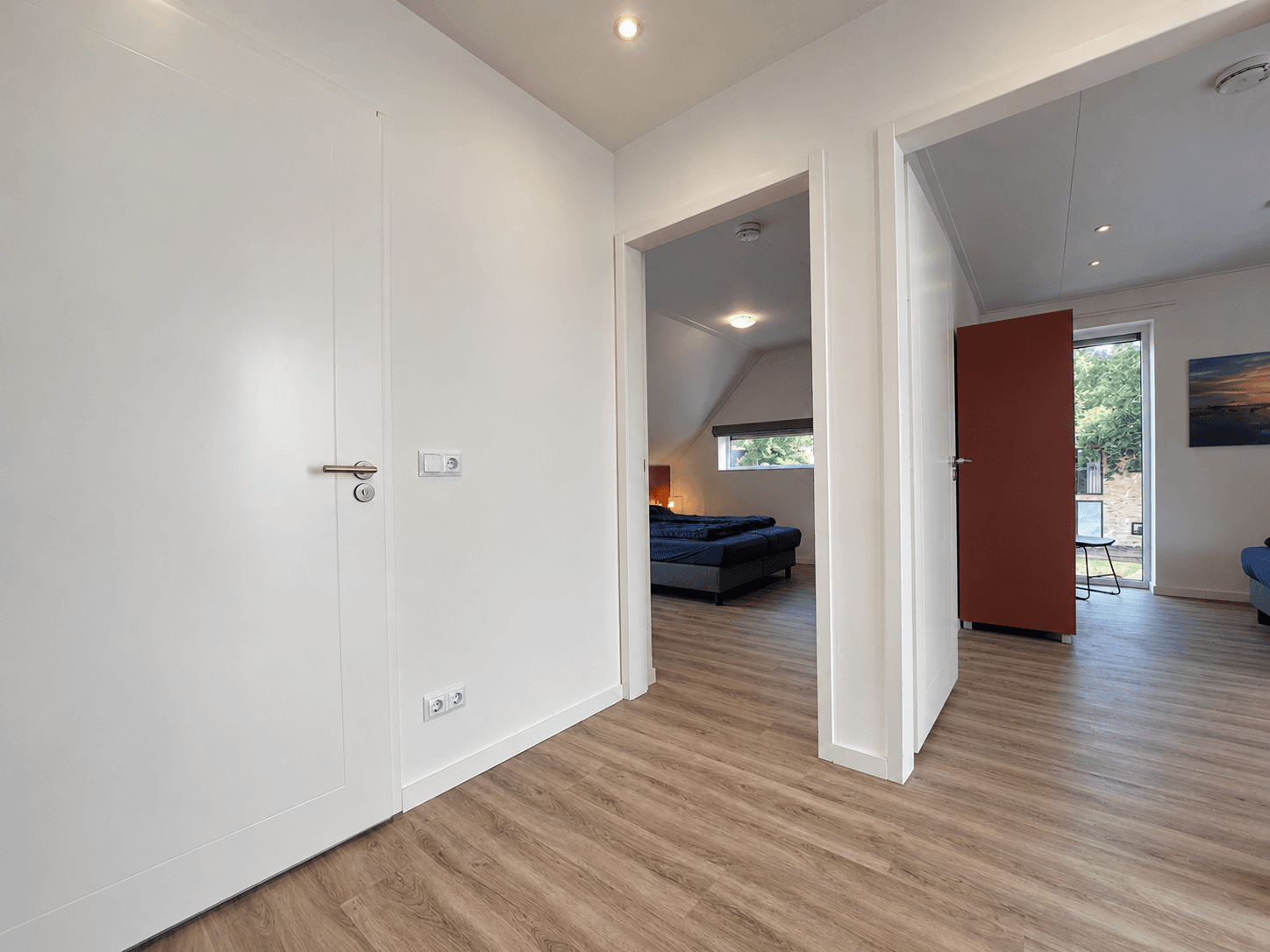 Ingang naar recreatiewoning met zicht op een woonkamer en slaapkamer, lichte wanden en houten vloer.