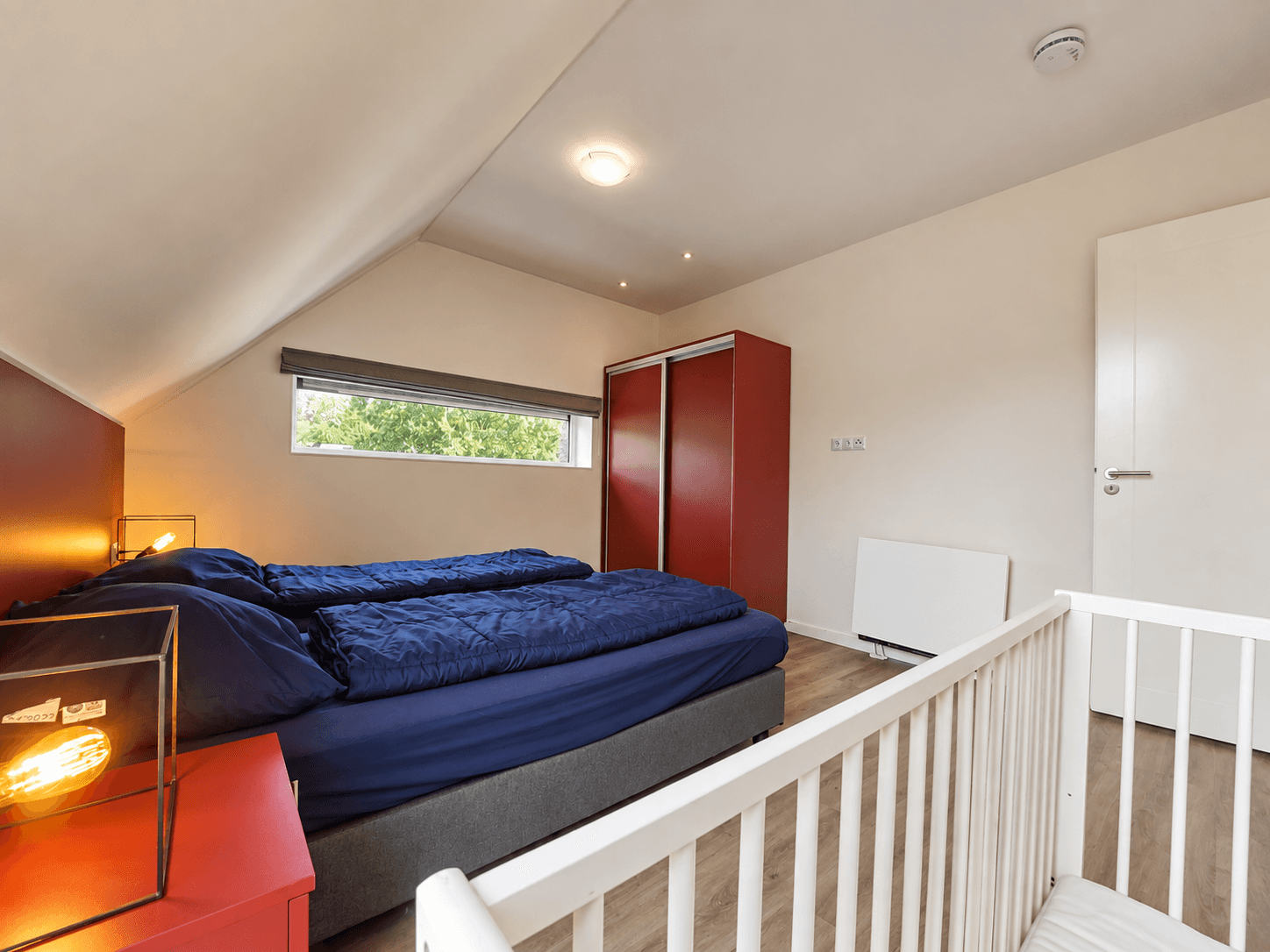 Slaapkamer met een tweepersoonsbed, kast en babybedje, lichte muren en raam met uitzicht op groen.