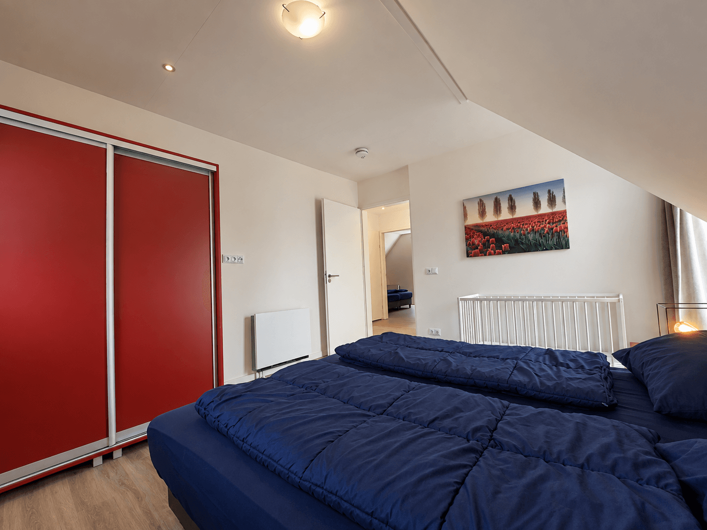Slaapkamer met twee eenpersoonsbedden, lichte wanden en een grote rode kast naast een schilderij van bloemen.