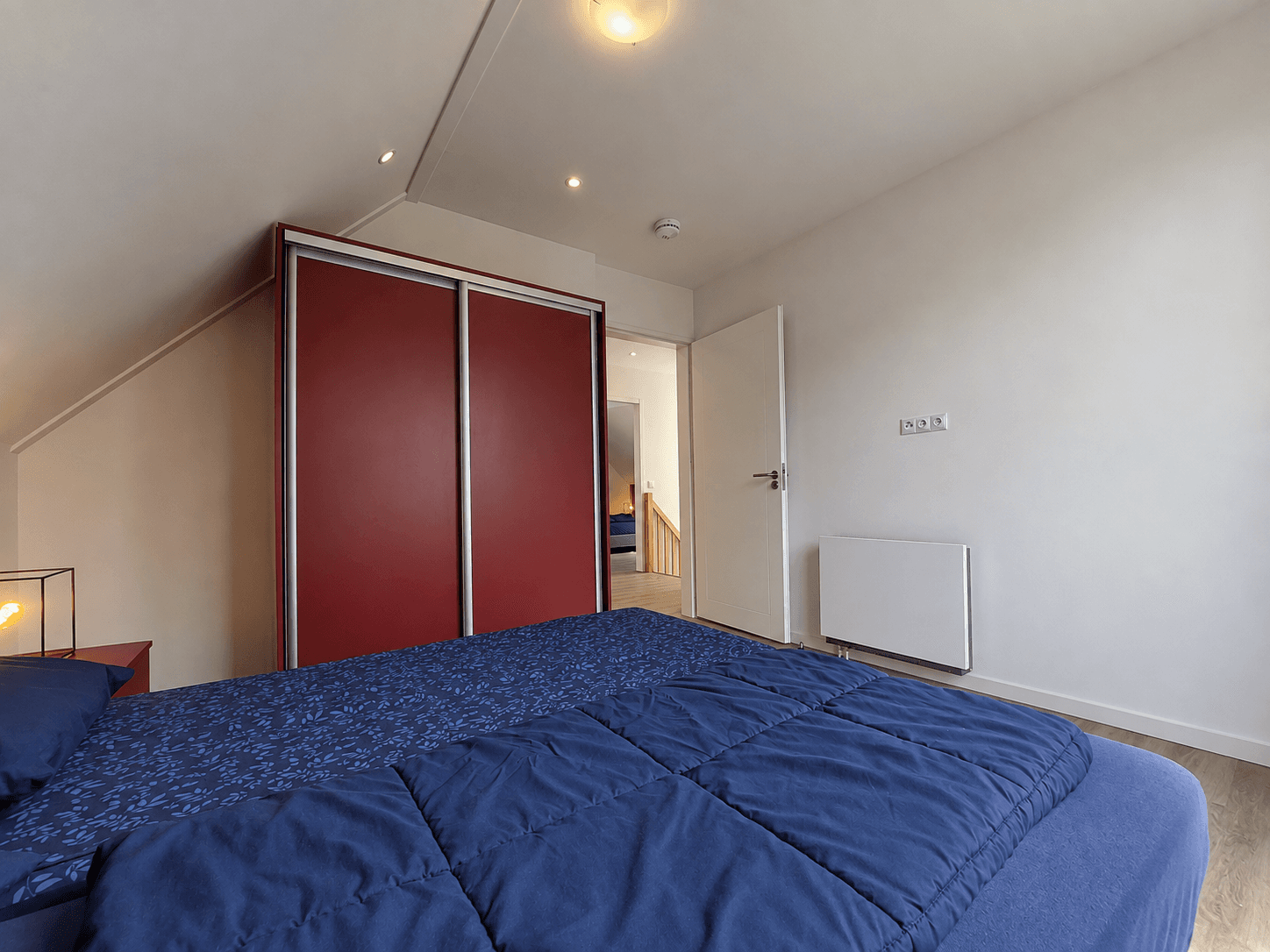 Slaapkamer met een bed, kast in rood en neutrale muren, toegang tot een aanpalende ruimte zichtbaar.