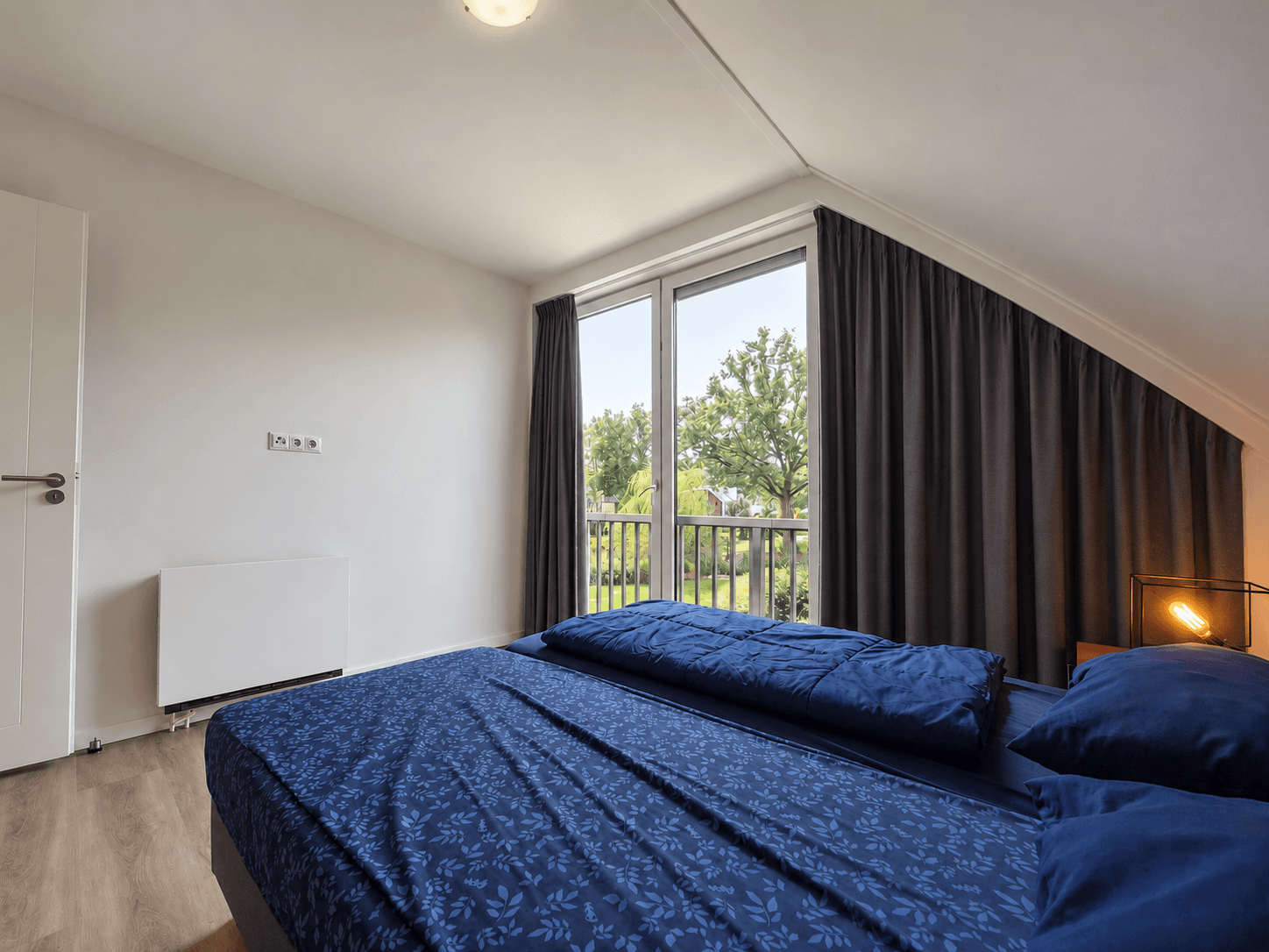 Slaapkamer met blauw beddengoed, opvallende ramen en grijze gordijnen zonder zicht op de buitenomgeving