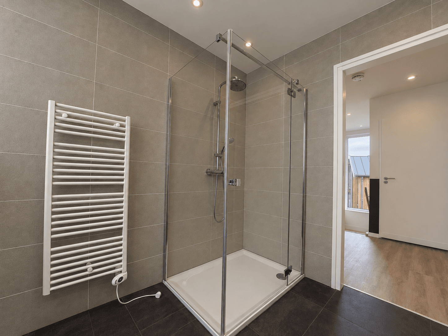 Douchecabine met glazen wanden, betegelde muren en een radiator in een ruime badkamer met houten vloer.
