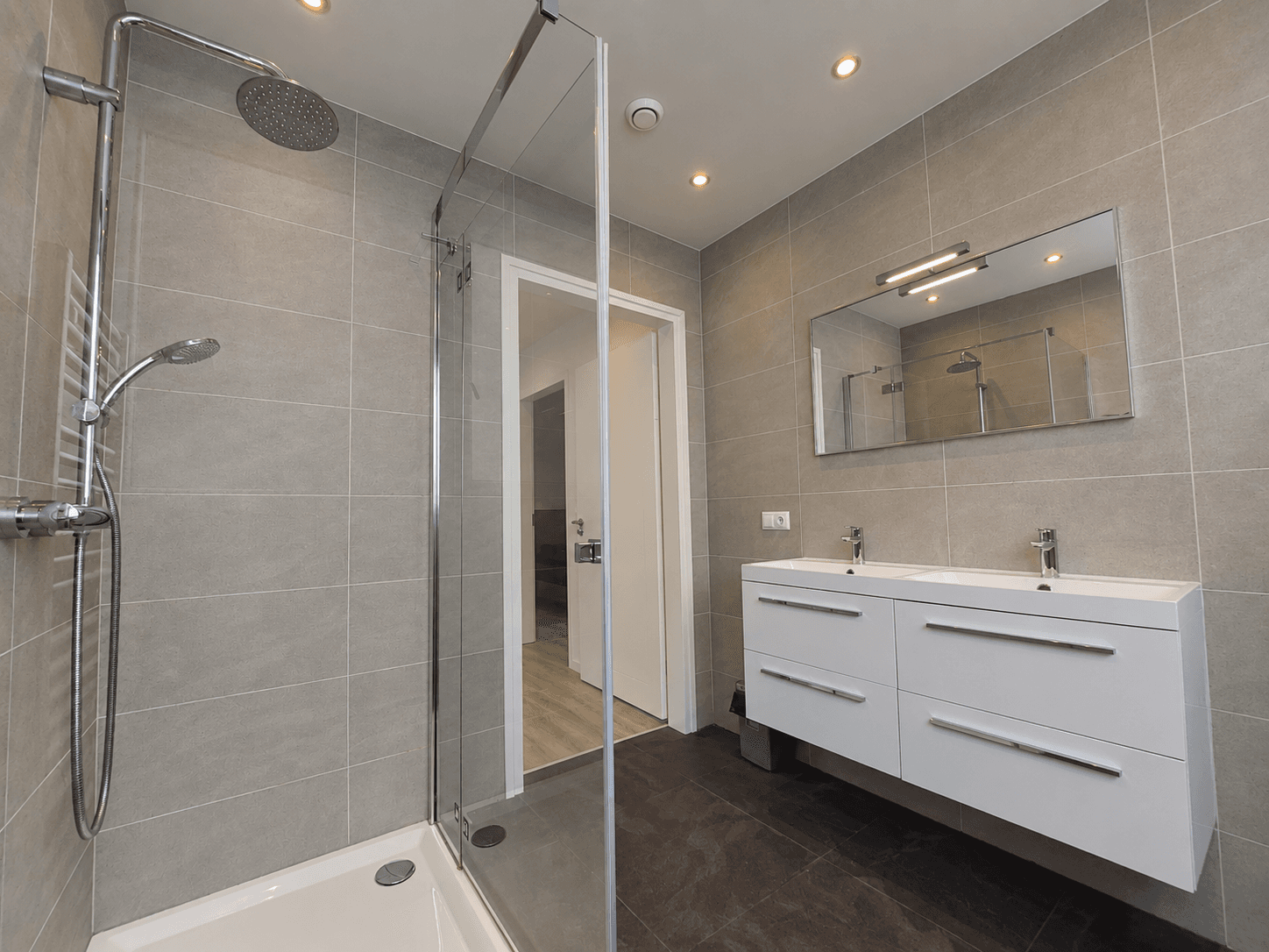 Moderne badkamer met inloopdouche, dubbele wastafel en grote spiegel, grijze wand- en vloertegels.