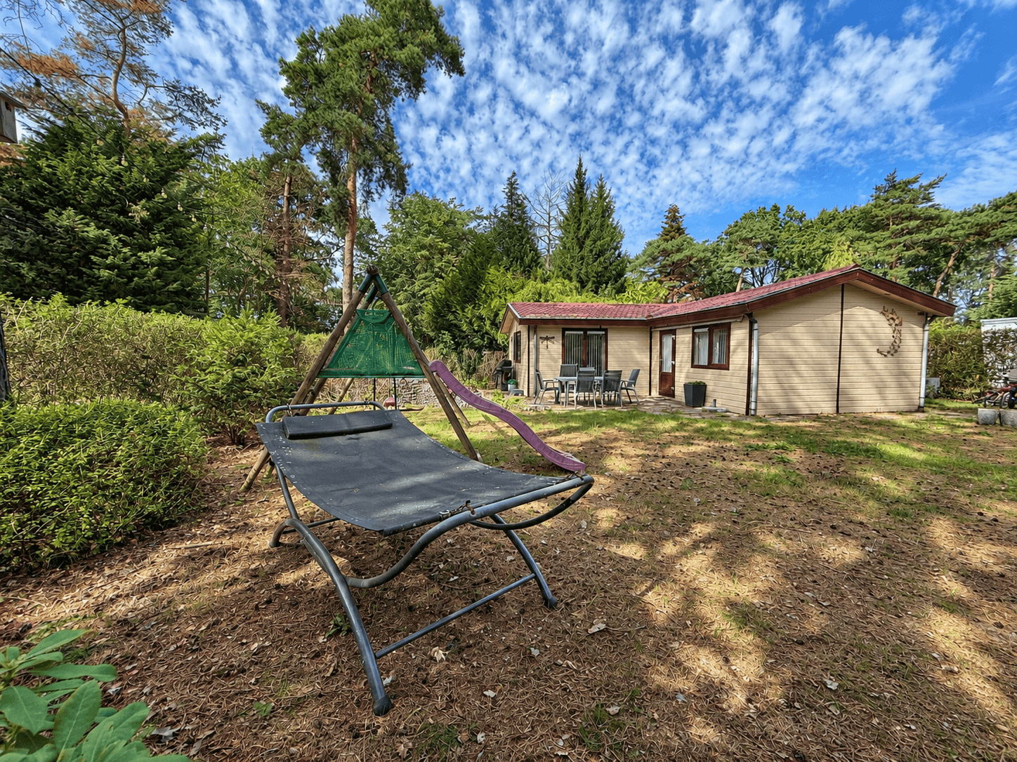 Recreatiewoning met groene tuin, hangstoel en terras, omringd door bomen en een heldere lucht.