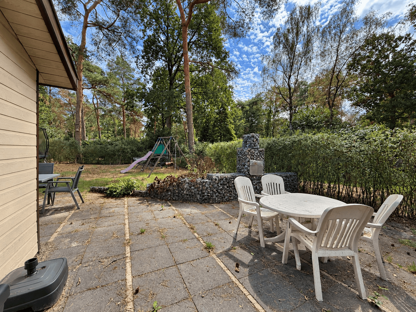 Terras met tuinmeubilair, speeltoestel en grill aan de zijkant van de recreatiewoning omringd door groen en bomen