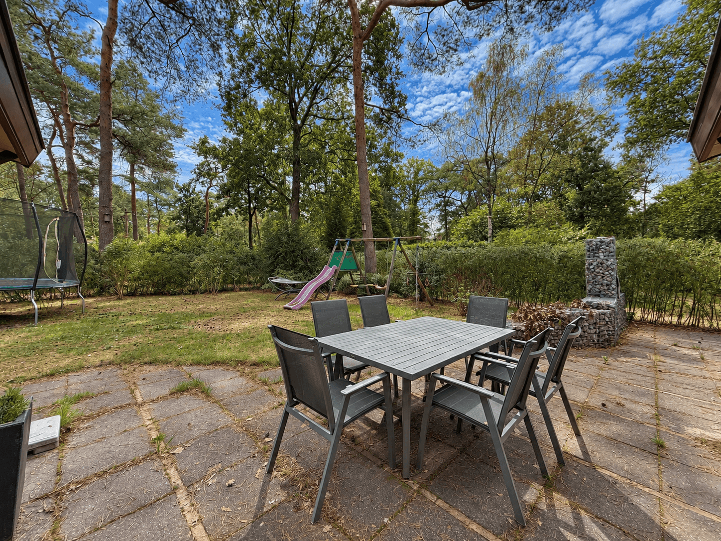 Terras met een tafel en stoelen, omringd door groen, met speeltoestellen en een trampoline op de achtergrond