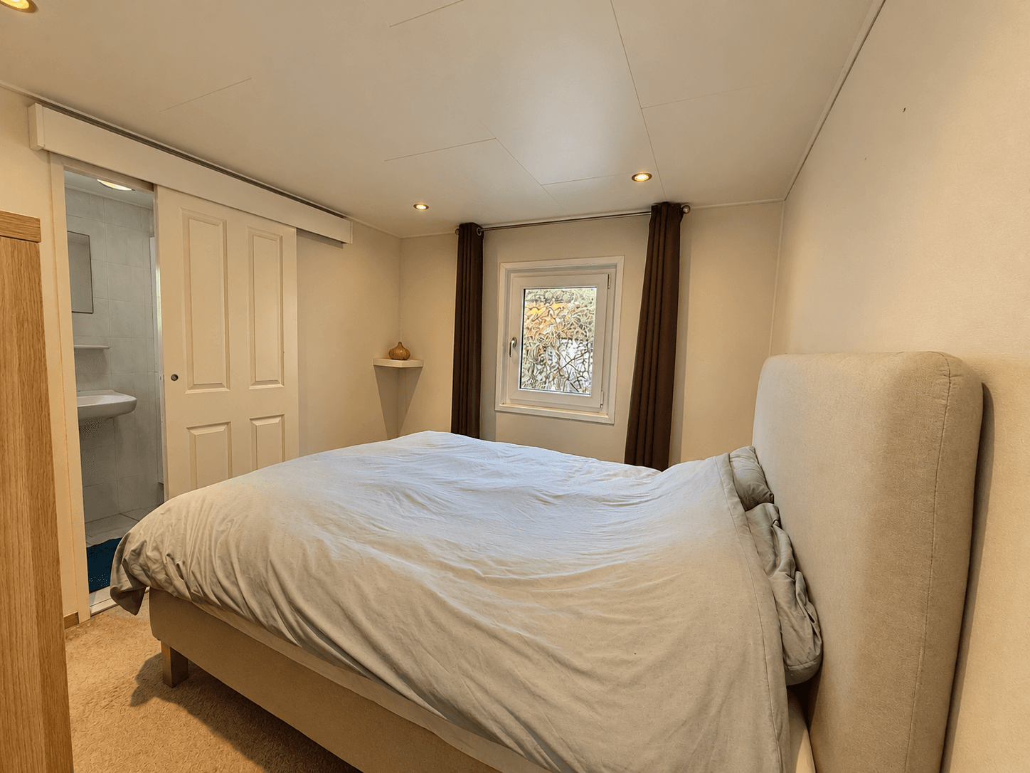 Sfeervolle slaapkamer met een bed, gordijnen en een raam naar buiten, badkamerdeur aan de achterzijde zichtbaar