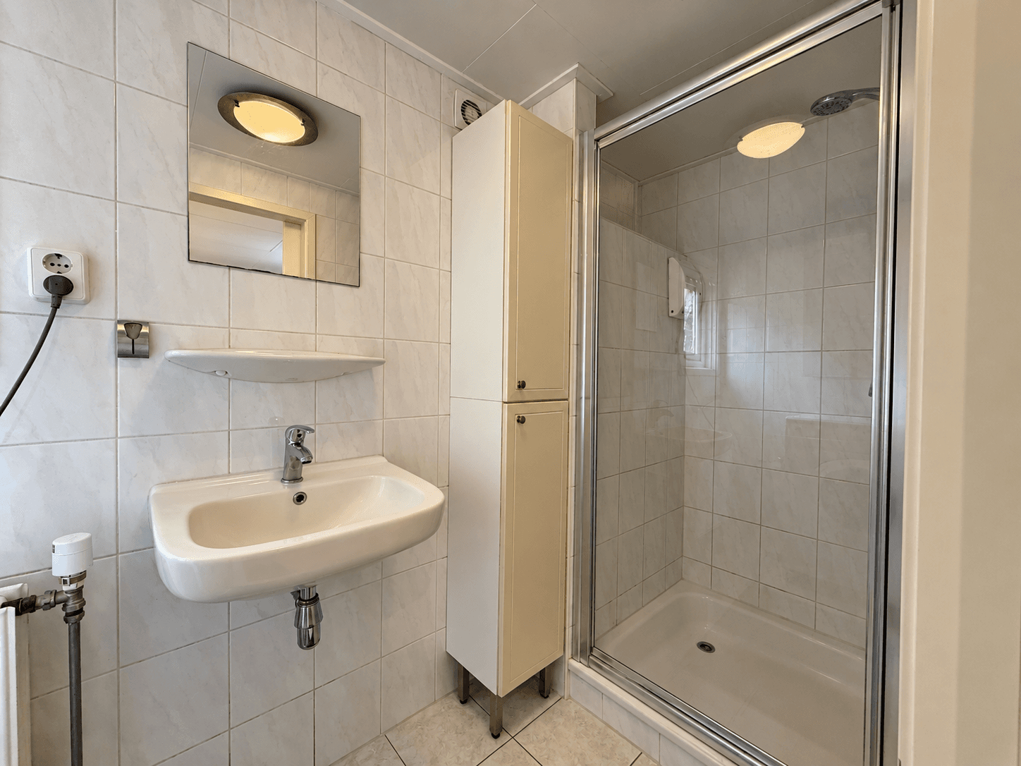 Badkamer met douchecabine, wastafel en opbergkast met wand- en vloertegels in lichte kleuren.