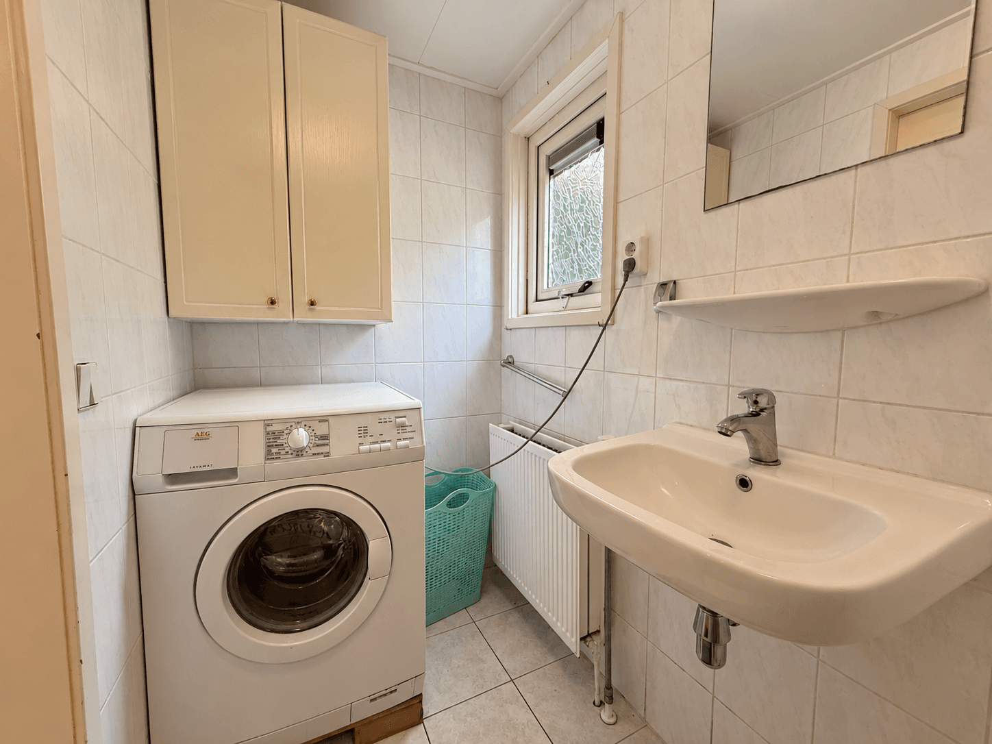 Wasmachine en wastafel in een lichte, betegelde ruimte met opbergkasten en een raam voor natuurlijk licht