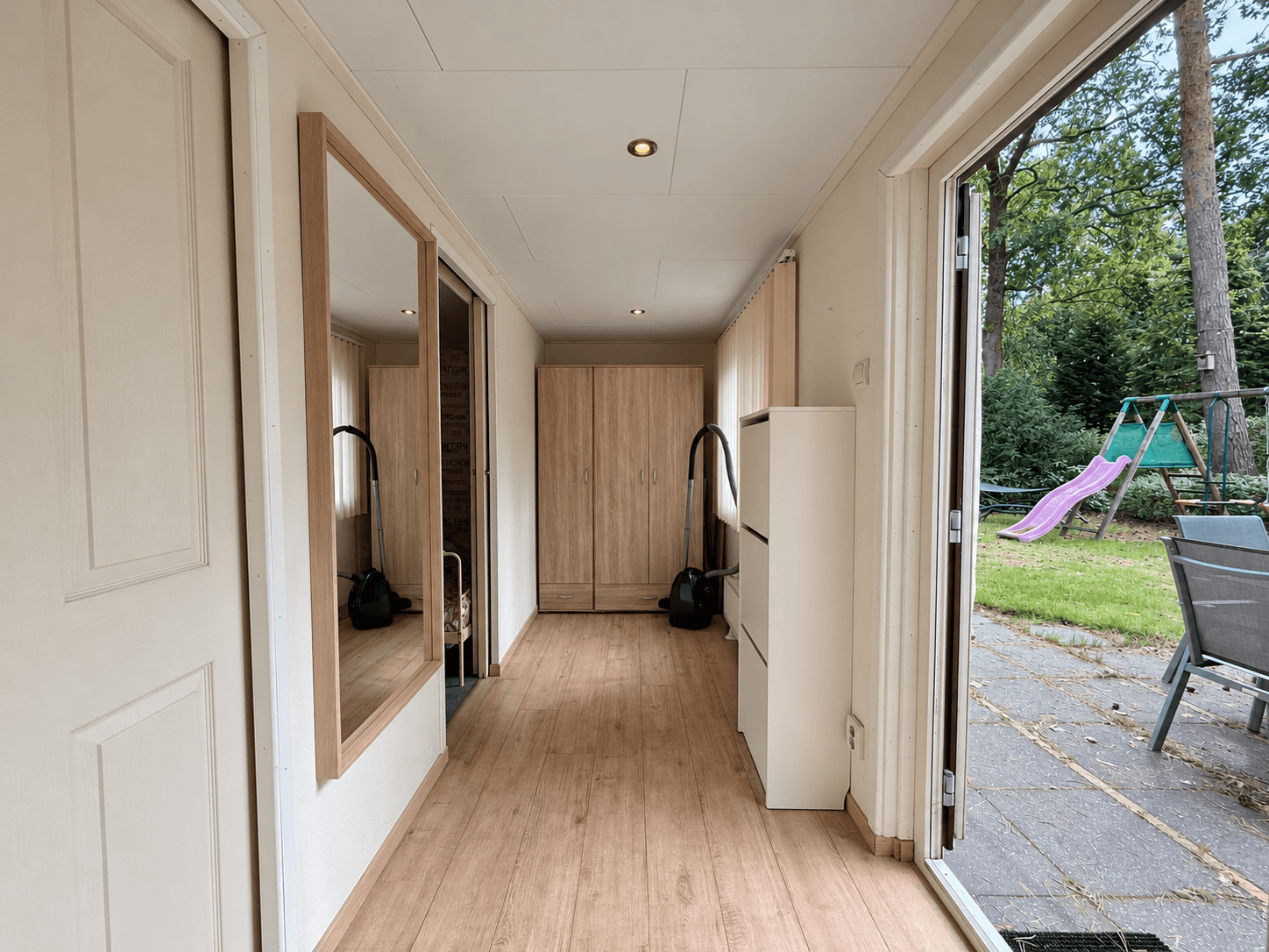 Entree van een recreatiewoning met een grote spiegel en kast, uitzicht op een tuin met schommel en speeltoestel buiten.