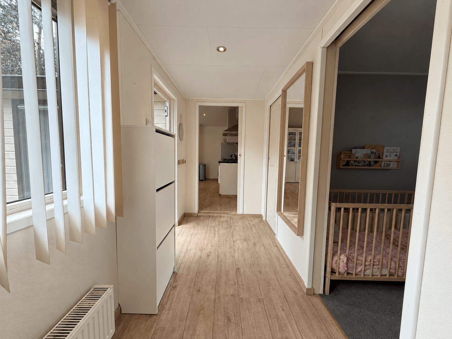 Lange gang met laminaatvloer, aan de rechterkant een slaapkamer met een ledikant, aan het einde zichtbaar een keuken.