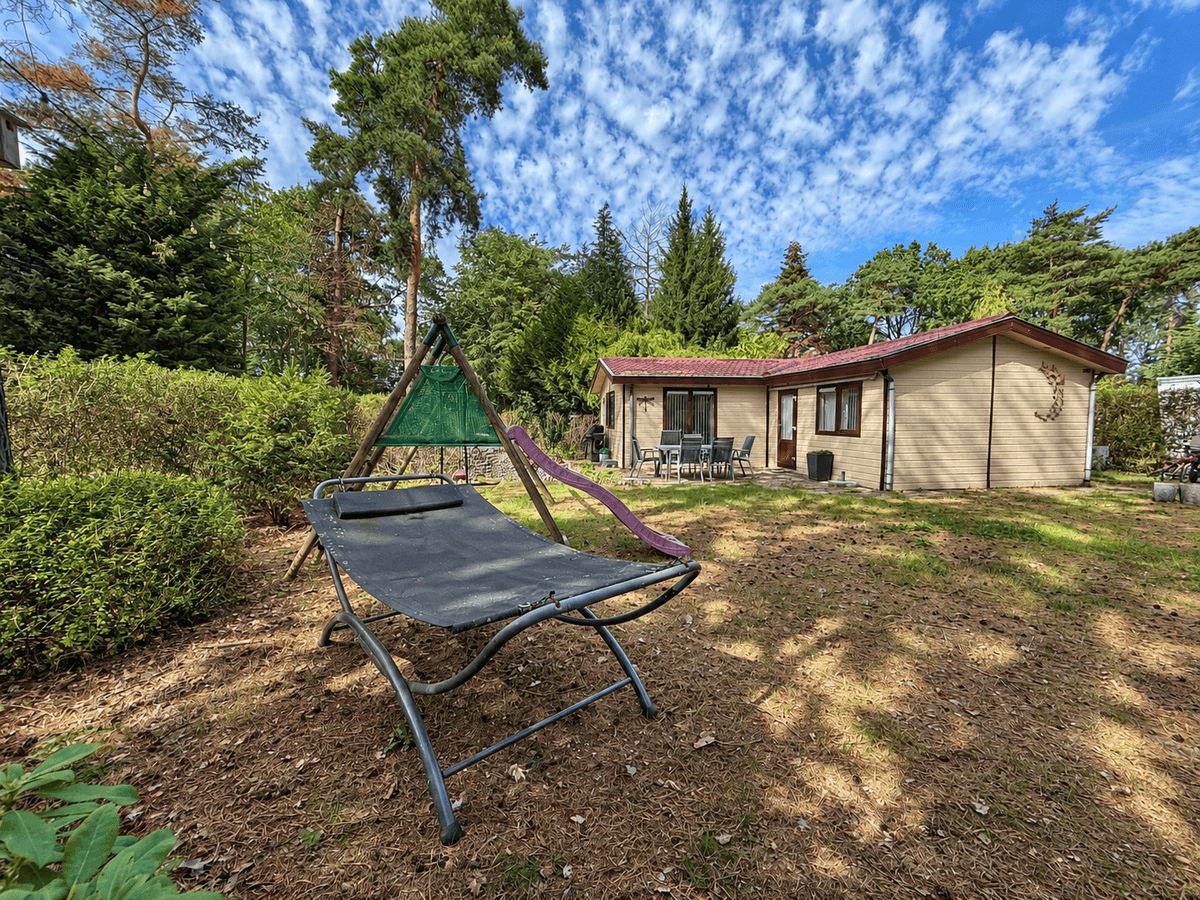 Recreatiewoning met groene tuin, hangstoel en terras, omringd door bomen en een heldere lucht.