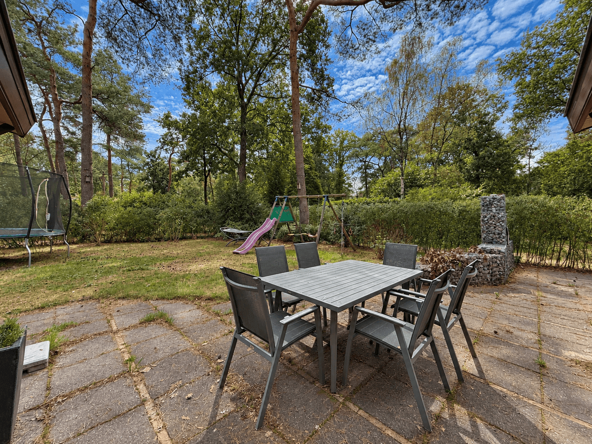 Terras met een tafel en stoelen, omringd door groen, met speeltoestellen en een trampoline op de achtergrond