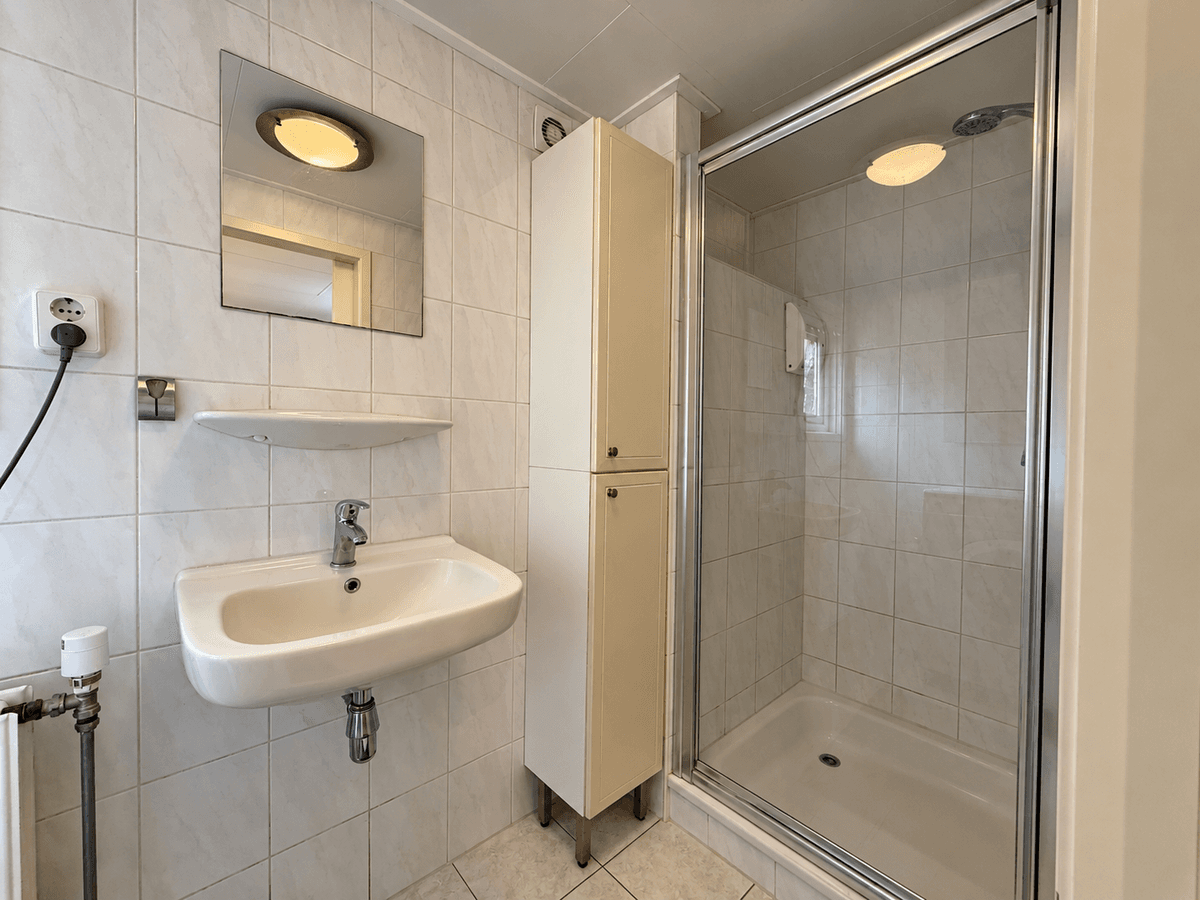 Badkamer met douchecabine, wastafel en opbergkast met wand- en vloertegels in lichte kleuren.
