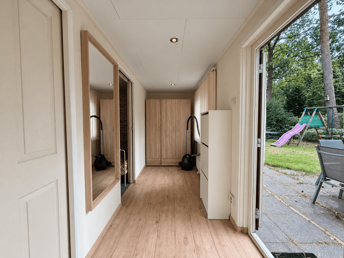 Entree van een recreatiewoning met een grote spiegel en kast, uitzicht op een tuin met schommel en speeltoestel buiten.