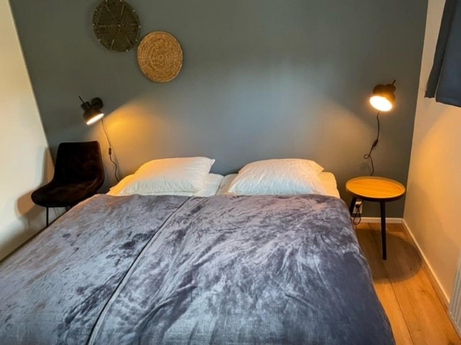 Slaapkamer met een tweepersoonsbed, nachtkastje, stoel en decoratieve wandobjecten op een grijze muur.