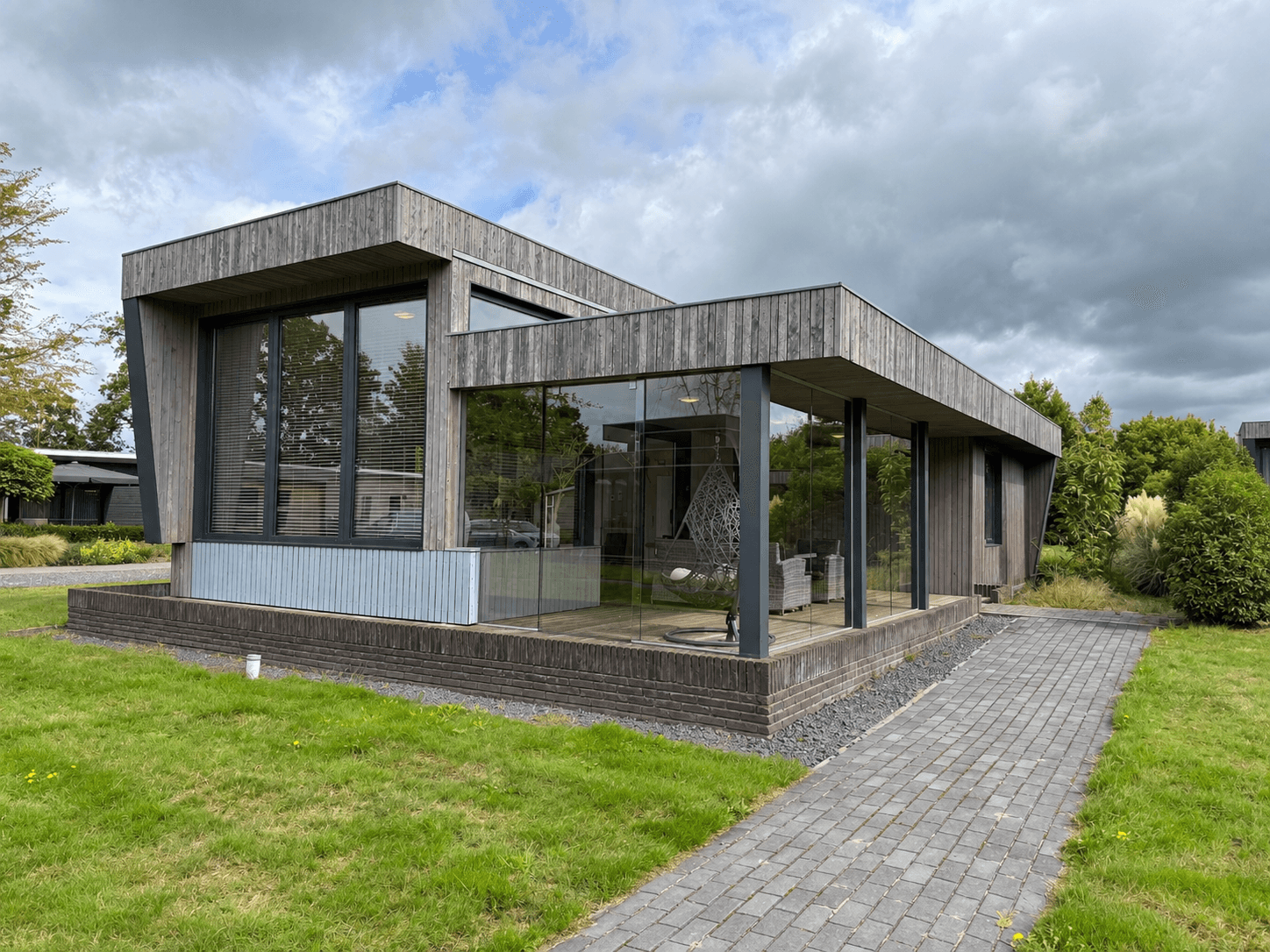 Moderne recreatiewoning met grote glazen wanden en houten façade, gelegen op een groen terrein met een betegeld pad