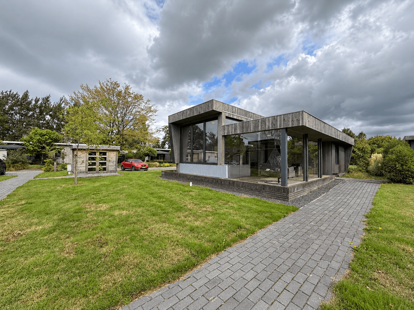 Moderne recreatiewoning met grote ramen en een uitgestrekt grasveld, omgeven door bomen en een oprit van klinkers.
