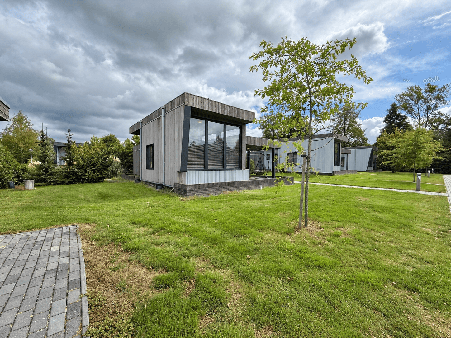 Moderne recreatiewoning met grote ramen, gelegen op een groen terrein met gras en een boom, aan een klinkerpad