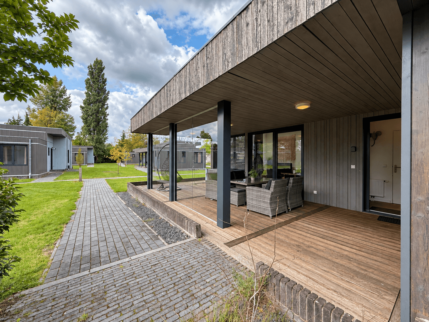 Moderne recreatiewoning met houten gevel, overdekt terras en zithoek, gelegen aan een groen grasveld met paden.
