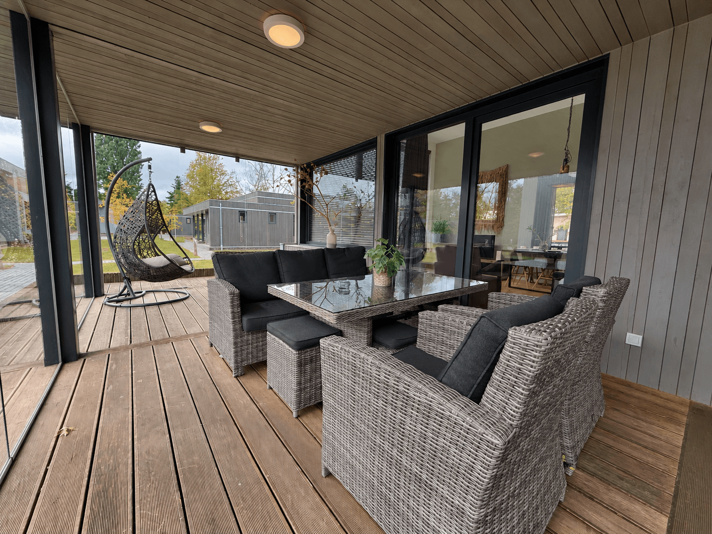 Overdekt terras met zithoek van wicker en glazen tafel, uitzicht op groene omgeving en vakantiehuisjes.