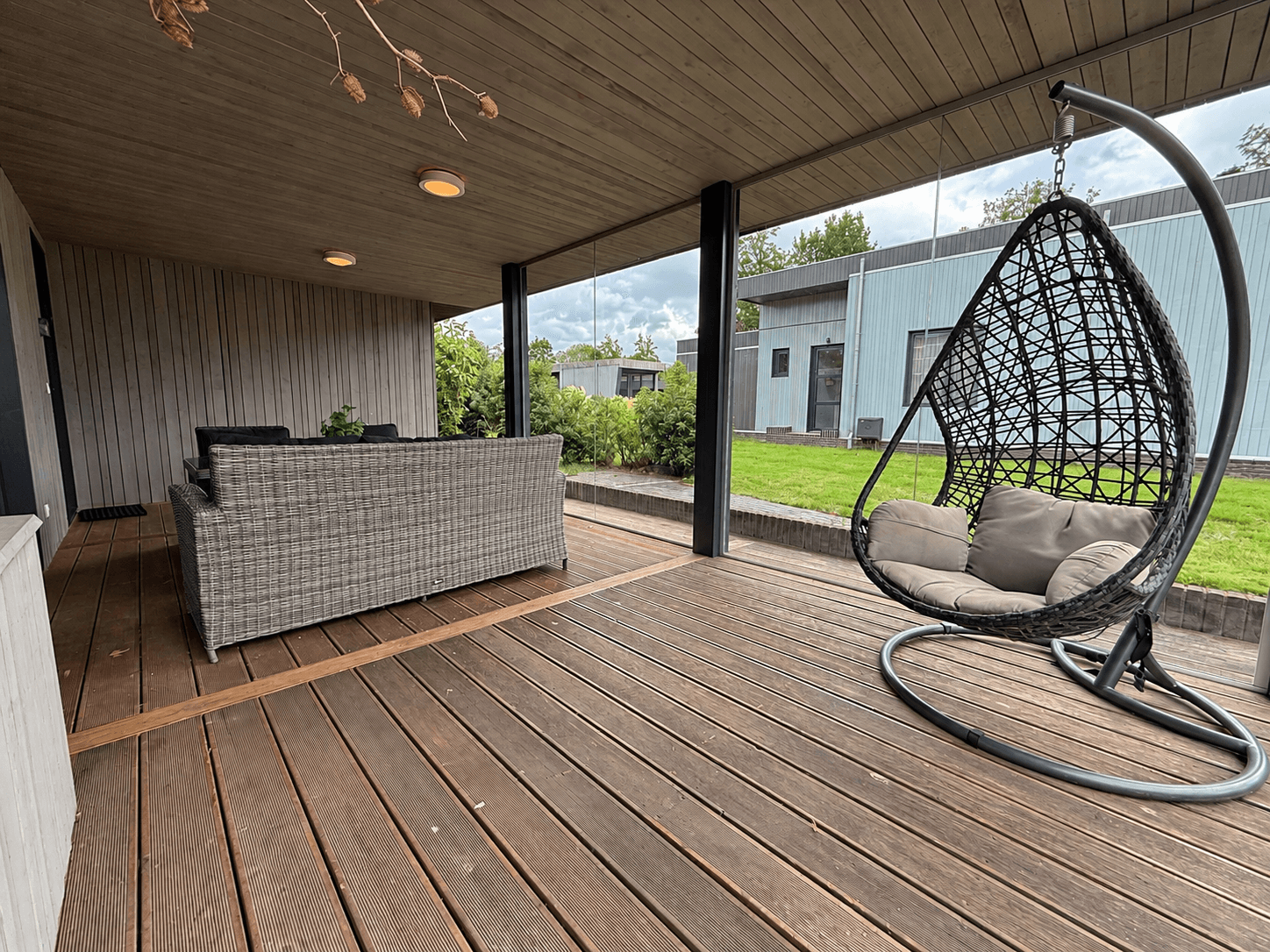 Overdekt terras met een hangstoel, een zithoek en zicht op het groene grasveld en omliggende woningen