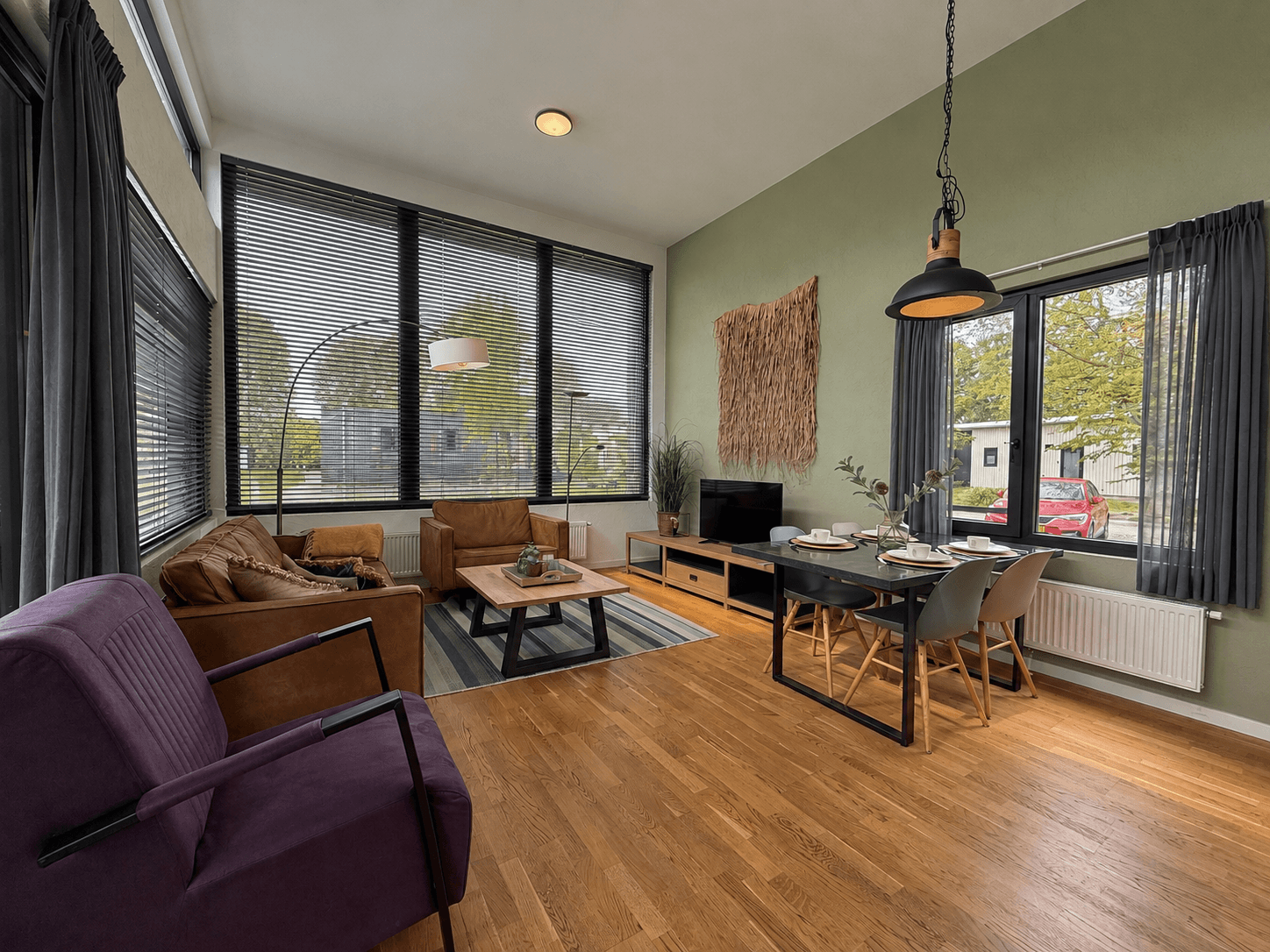 Ruime woonkamer met grote ramen, moderne meubels en een eettafel, voorzien van een houten vloer en groene muur.