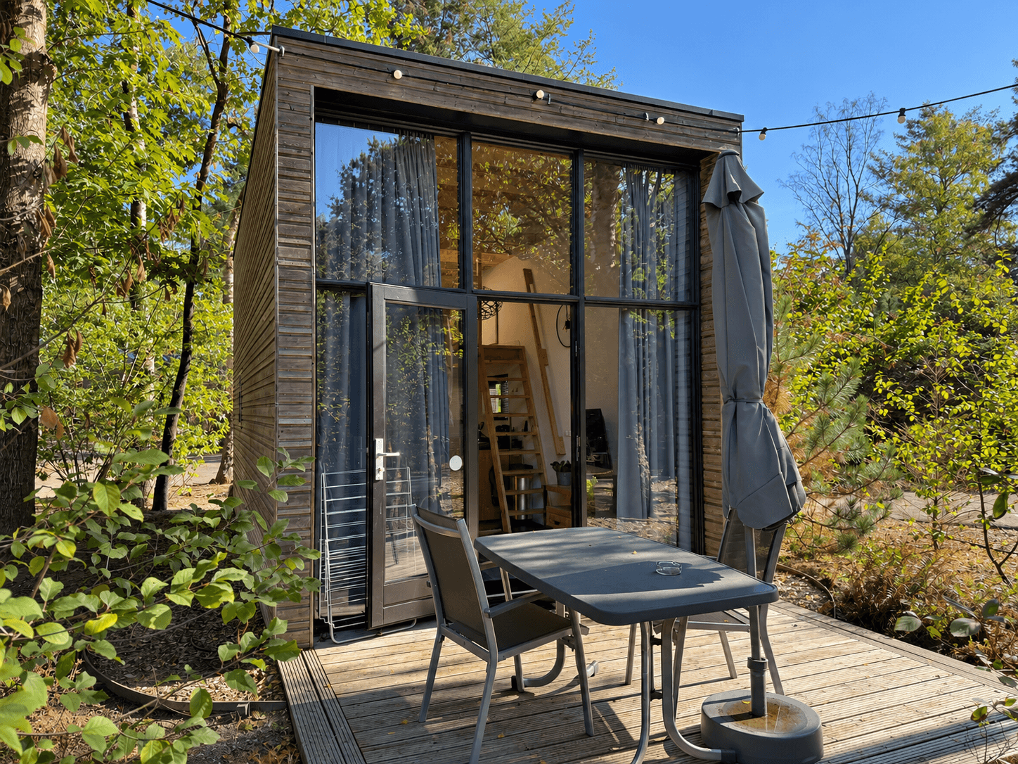Moderne recreatiewoning met grote raampartijen, terras met tafel en parasol, omringd door bomen en groen.