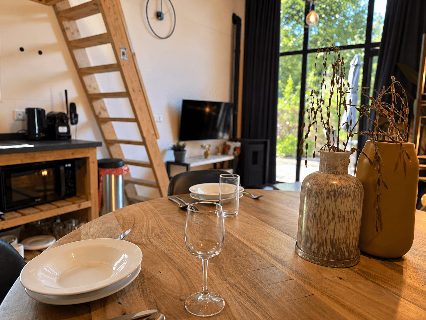 Eettafel met borden en glazen, open keuken met ladder naar bovenverdieping, grote ramen met uitzicht op bomen