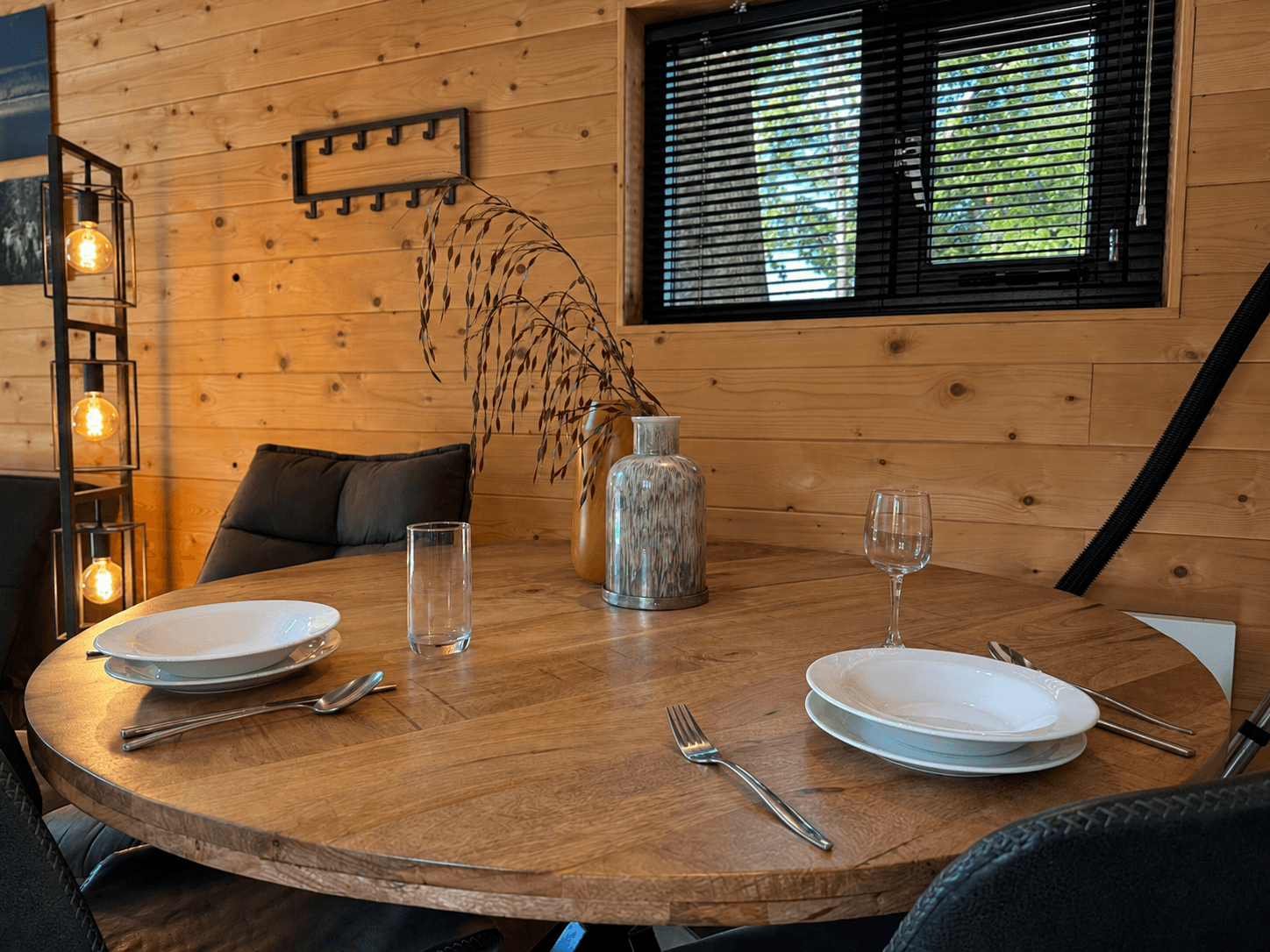 Eetkamertafel met ronde houten blad, gedekte tafel met borden en glazen, houten wand en raam met jaloezieën