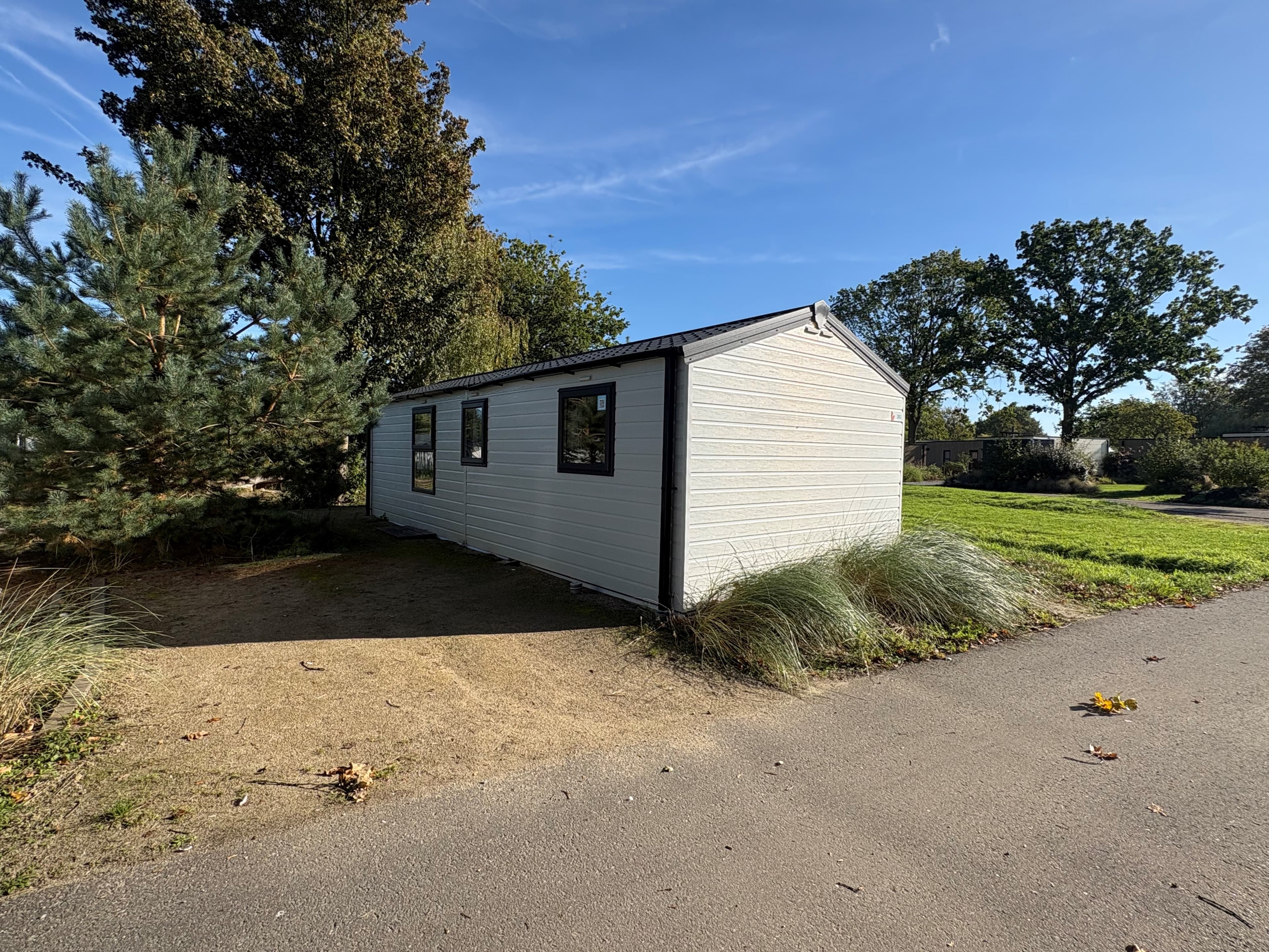 Recreatiewoning met lichtgekleurde gevel en enkele ramen, omringd door groen en een grindpad.