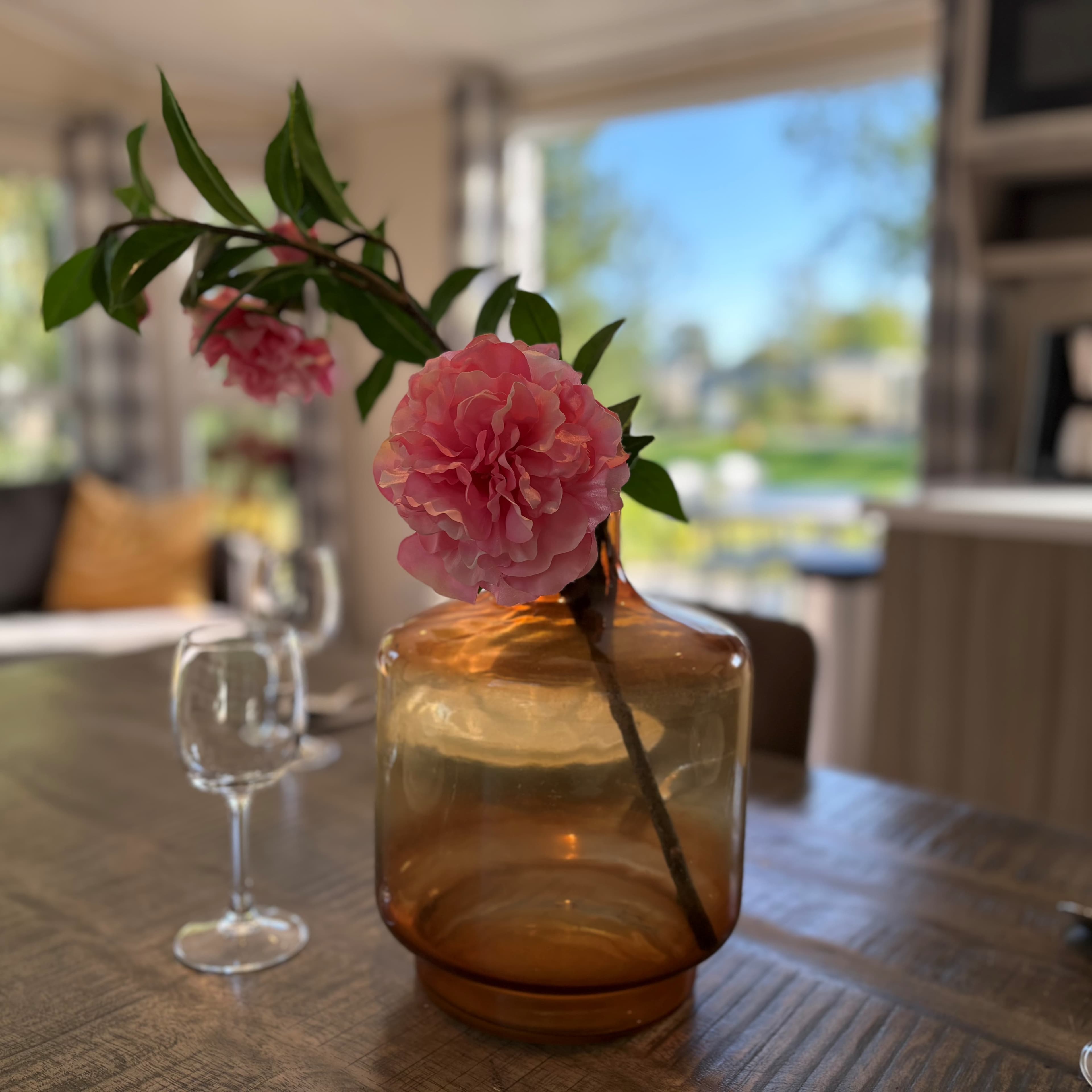 Een glazen vaas met een roze bloem staat op een houten tafel, op de achtergrond een zonnig uitzicht naar buiten.