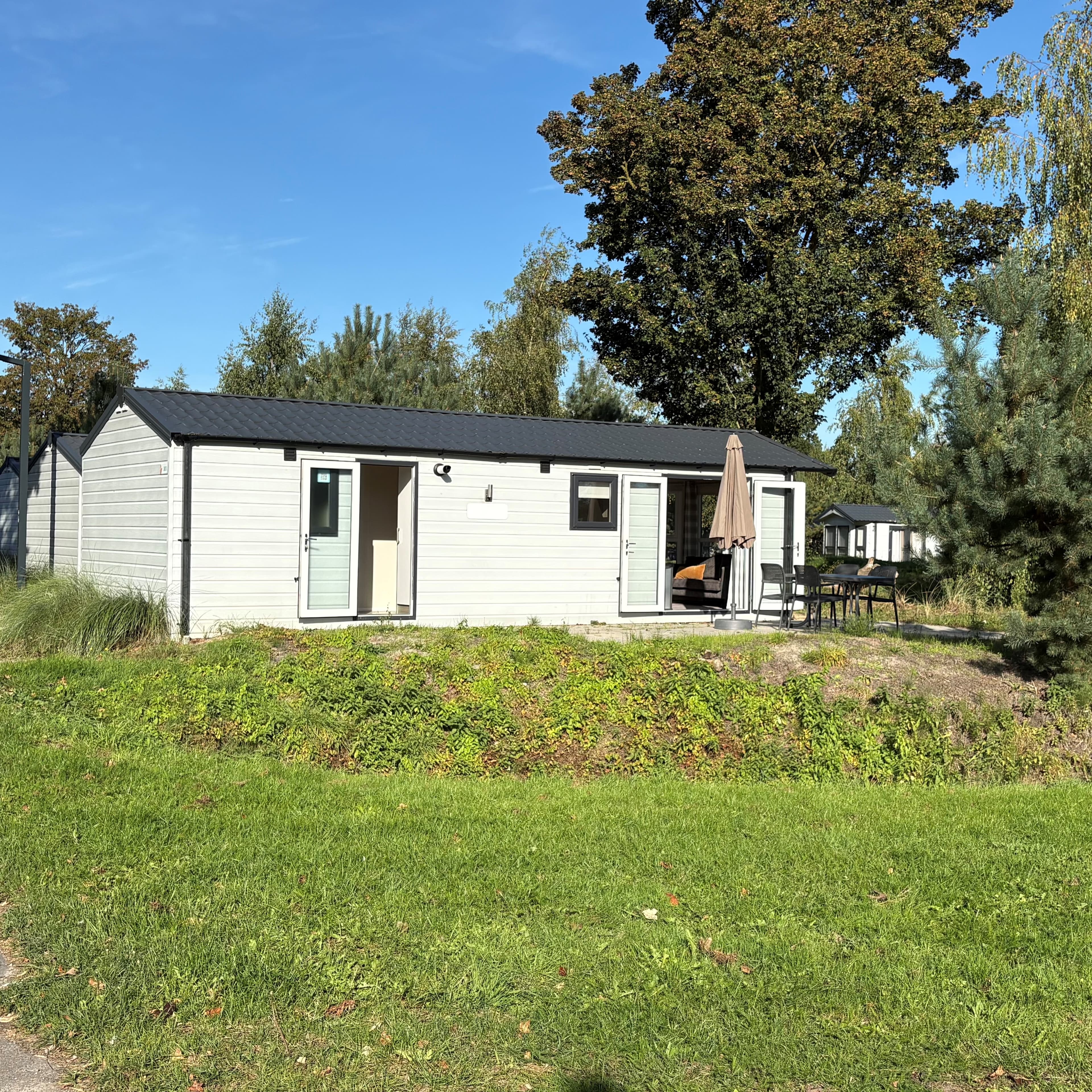 Houten recreatiewoning met een terras en parasol, omgeven door groen en bomen in een rustige omgeving