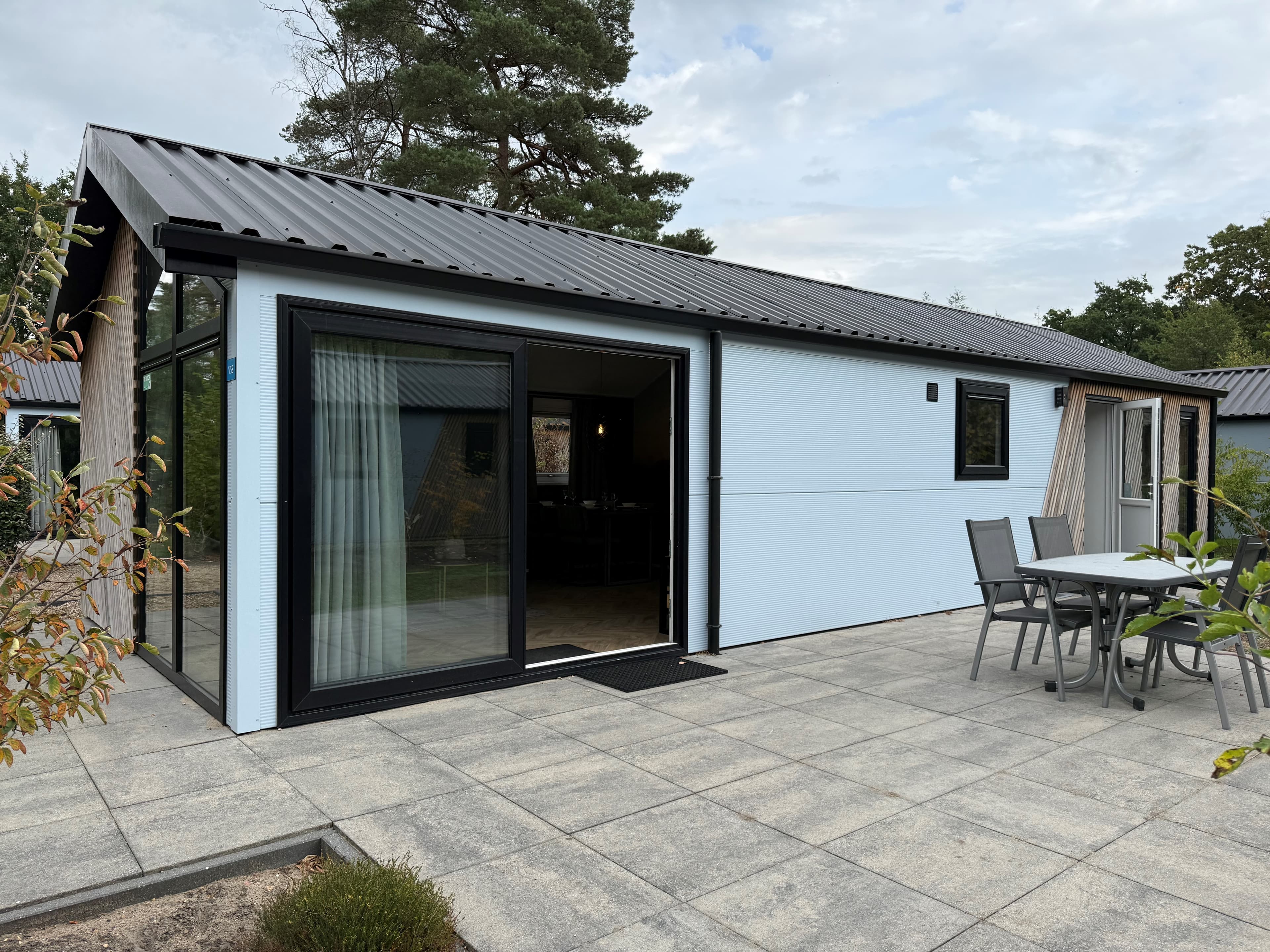 Moderne recreatiewoning met een terras en tuinmeubilair, gelegen in een groene omgeving met bomen en paden.