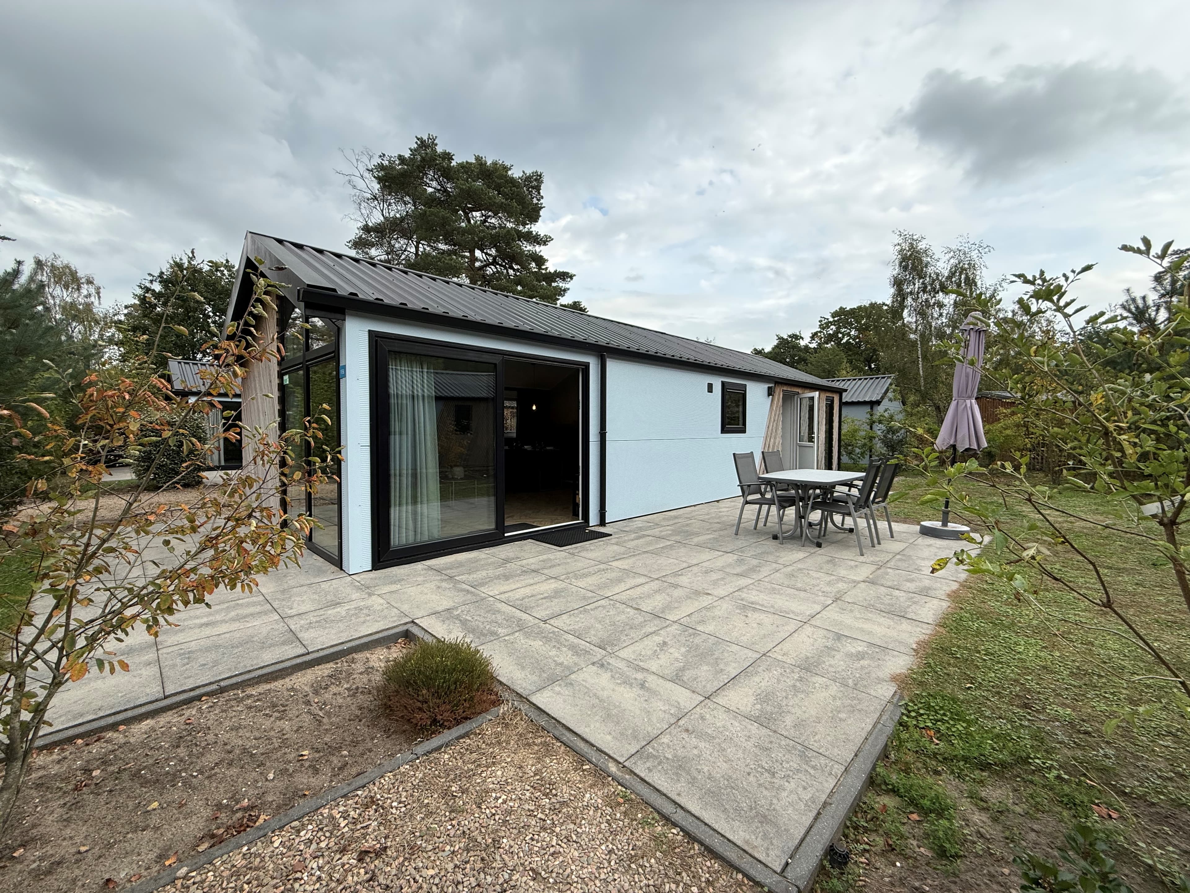 Moderne recreatiewoning met een terras, eettafel en parasol, omringd door groen en een betegeld pad.