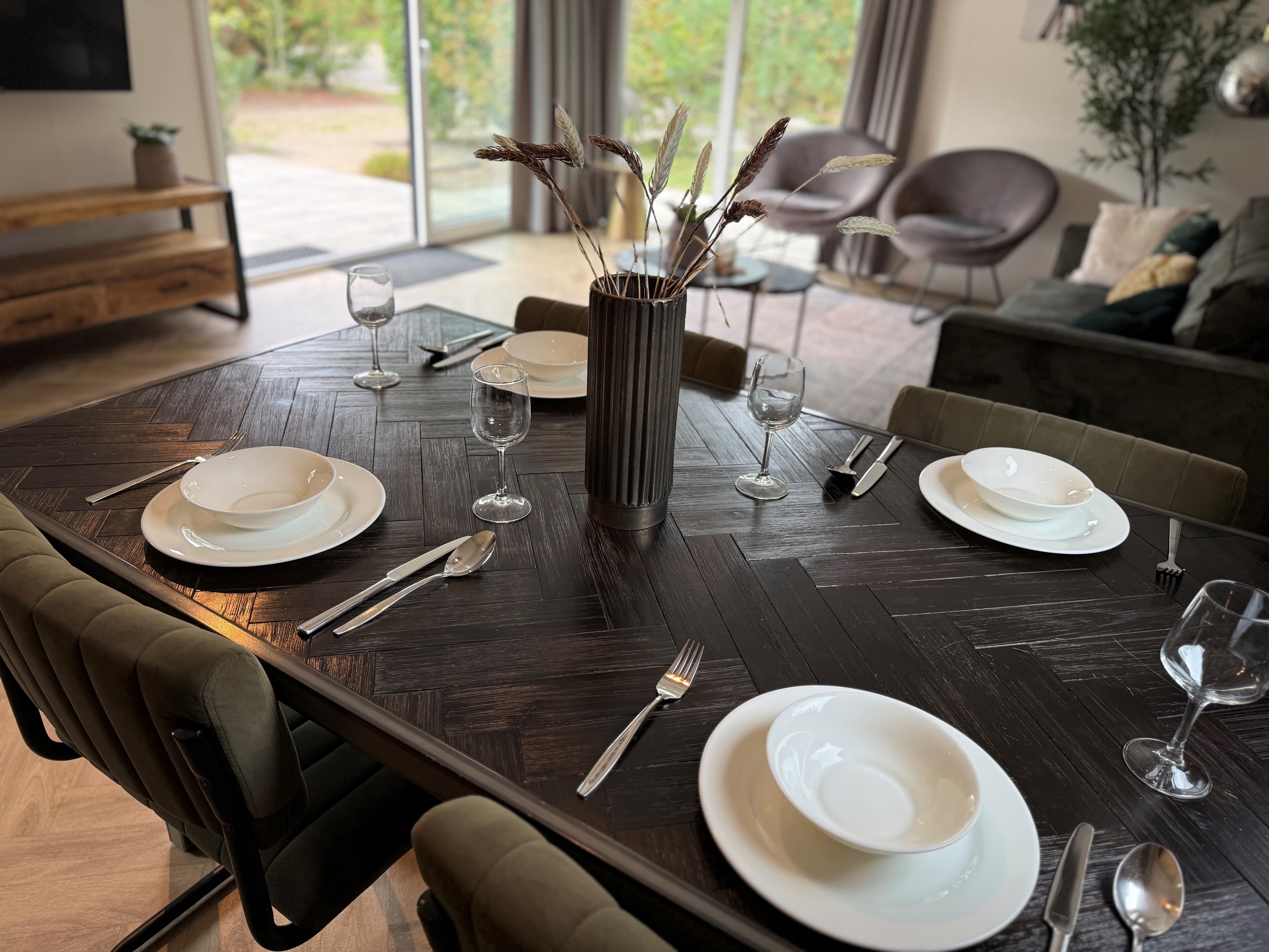 Eettafel met six setten borden, glazen en bestek, moderne stoelen en een vaas met takken in een woonkamer met grote ramen