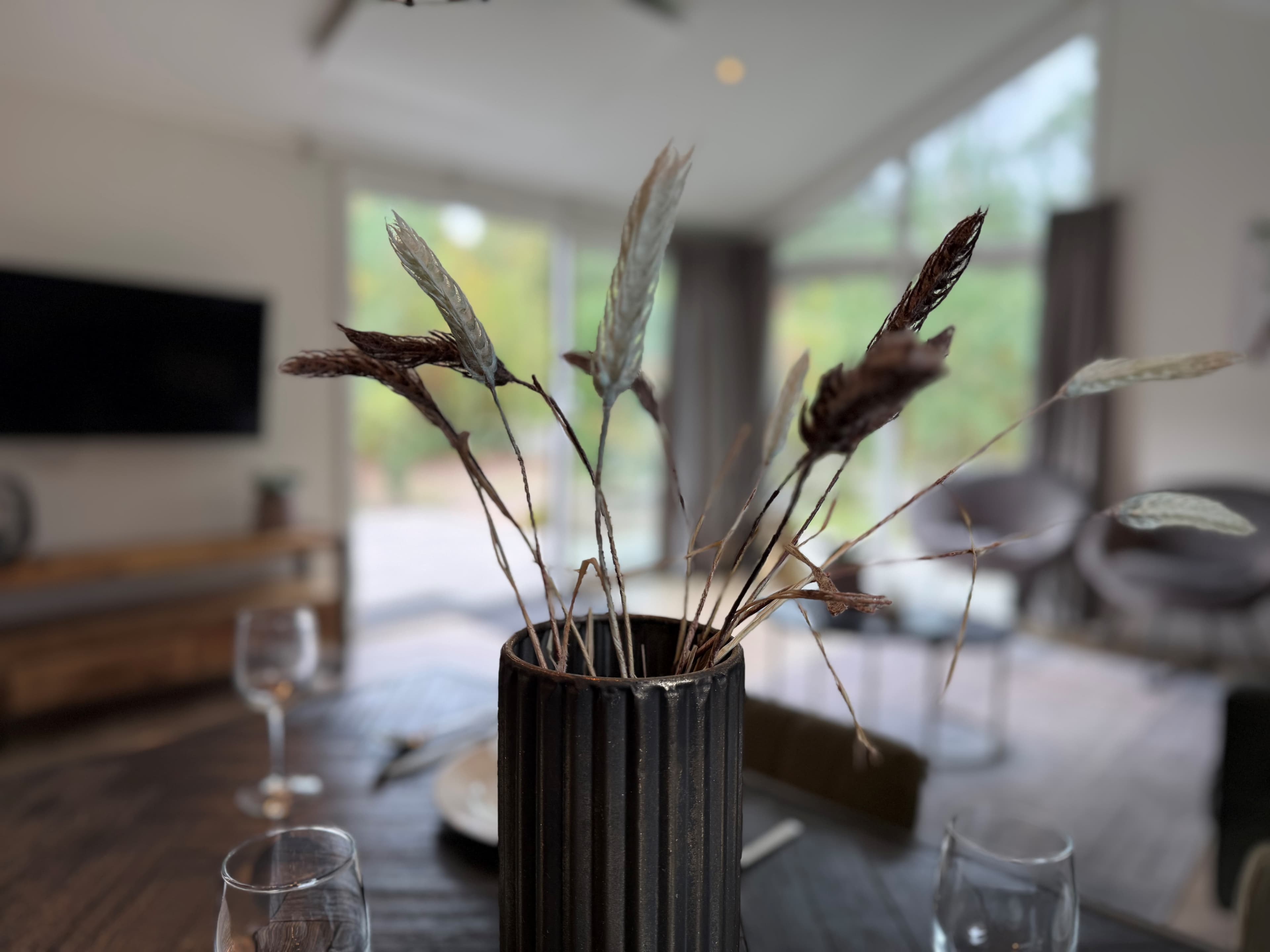 Interieur van een recreatiewoning met een vaas en droogbloemen op een houten tafel, grote ramen met uitzicht op de natuur